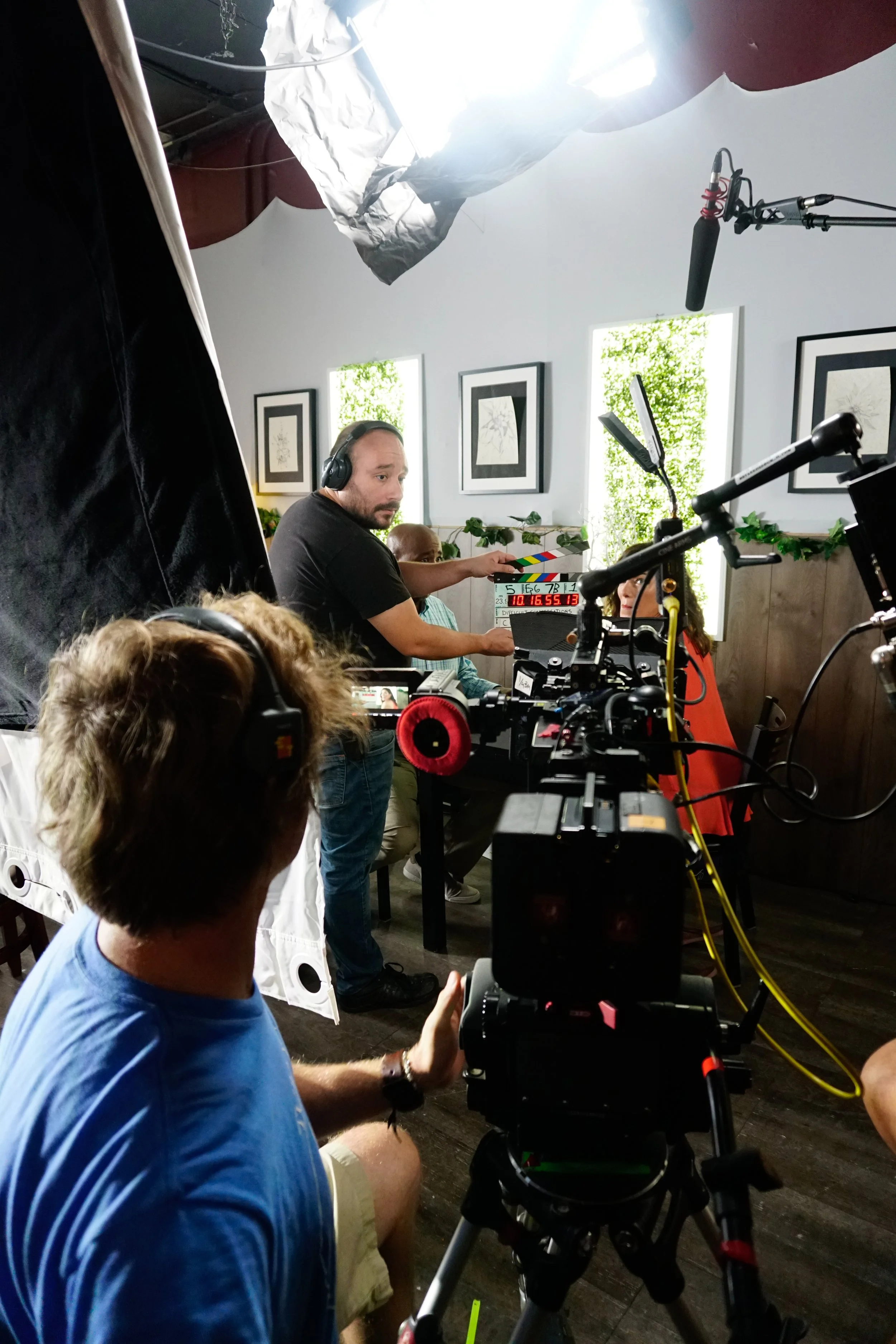 Filming crew on a set with professional camera and equipment, crew members working with actors in a well-lit room with framed artwork on the walls.