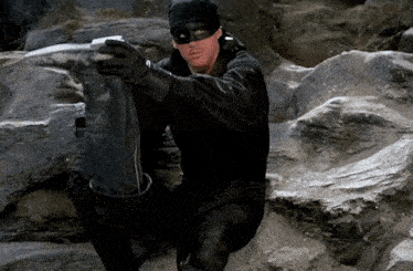 A person dressed in black, wearing a mask, crouching among rocks outdoors.