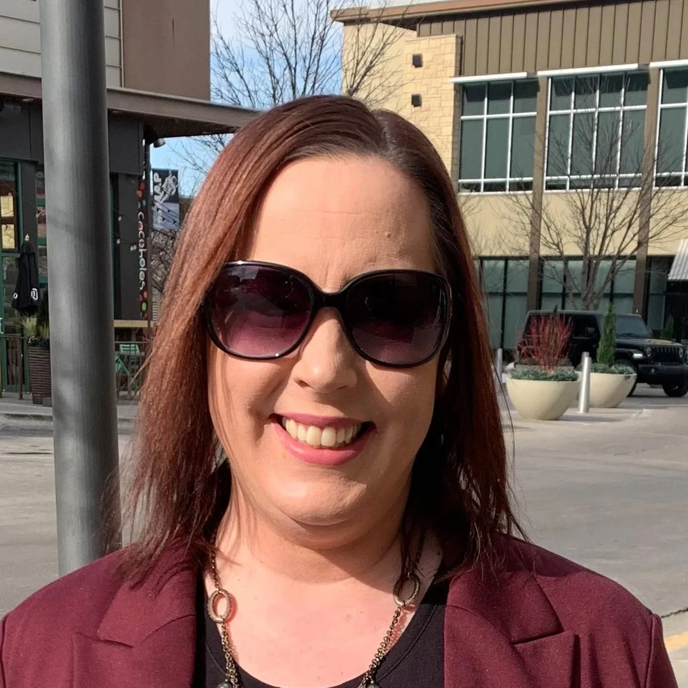 A woman with red hair wearing large sunglasses, a maroon blazer, and a black top, smiling outdoors on a sunny day.