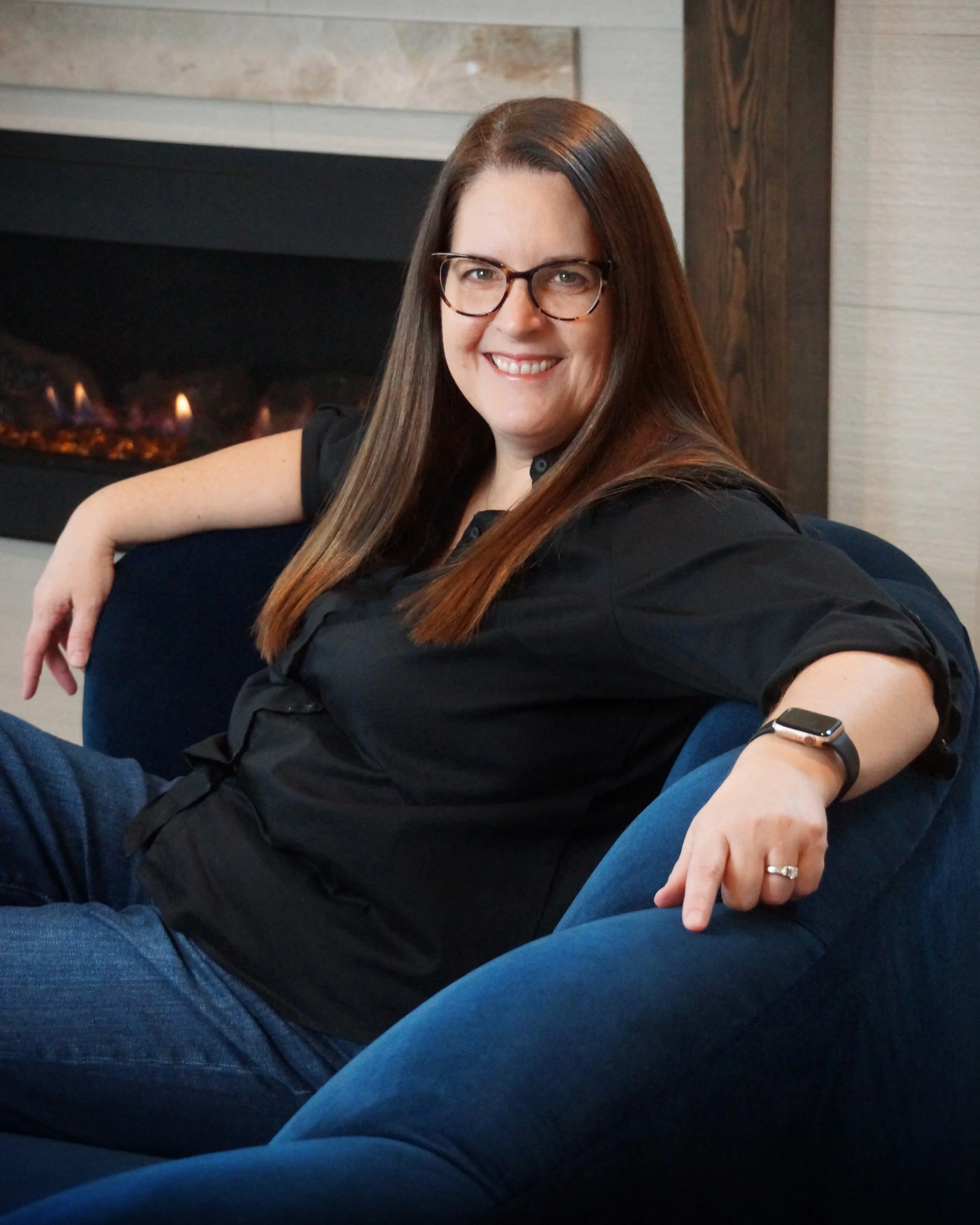 Woman with long brown hair, wearing glasses, black shirt, and a fitness tracker, sitting on a navy blue couch near a fireplace, smiling at the camera.