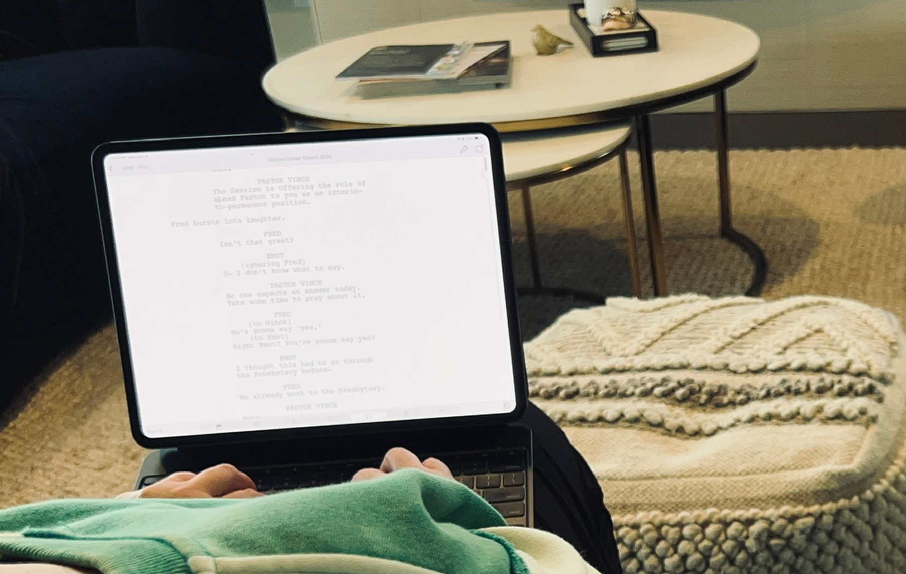 A cozy lounge area with two round white nesting tables, one with a small potted plant, a tray, and some magazines, a large fireplace in the background, and a blue armchair on the right. In the foreground, a person using a tablet or e-reader, with a patterned knitted blanket on their lap, on a beige woven carpet.