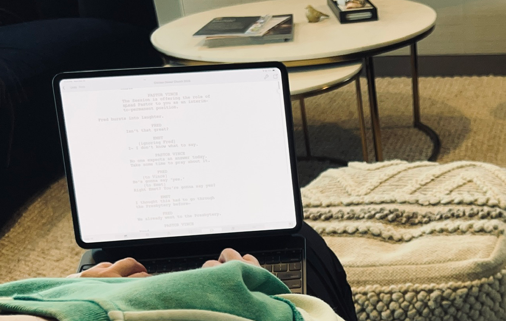 A cozy hotel room with a white oval coffee table, a decorative plant, a tray with a seashell, a magazine, and a fireplace in the background. In the foreground, a person is sitting with a laptop on their lap, displaying a script or text. To the right, there is a knitted ottoman and a blue armchair.