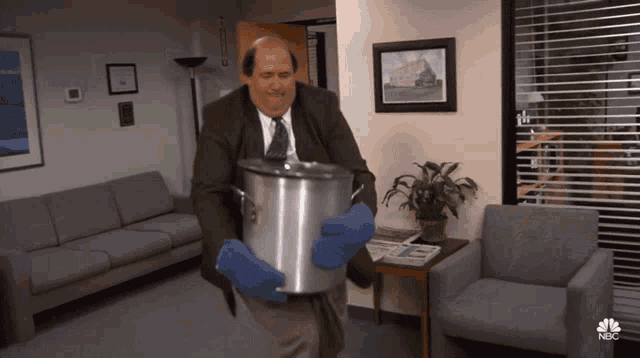 A man in a suit and blue oven mitts carrying a large metal pot in an office lounge area.