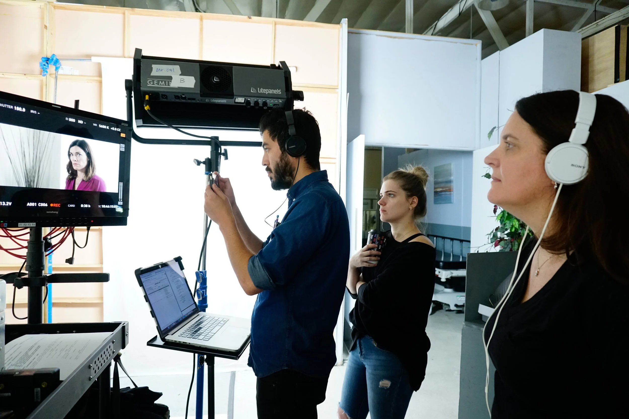 People working on a video shoot in a studio. A man is adjusting equipment, a woman holds a script, and another woman wears headphones and watches the scene on a monitor.