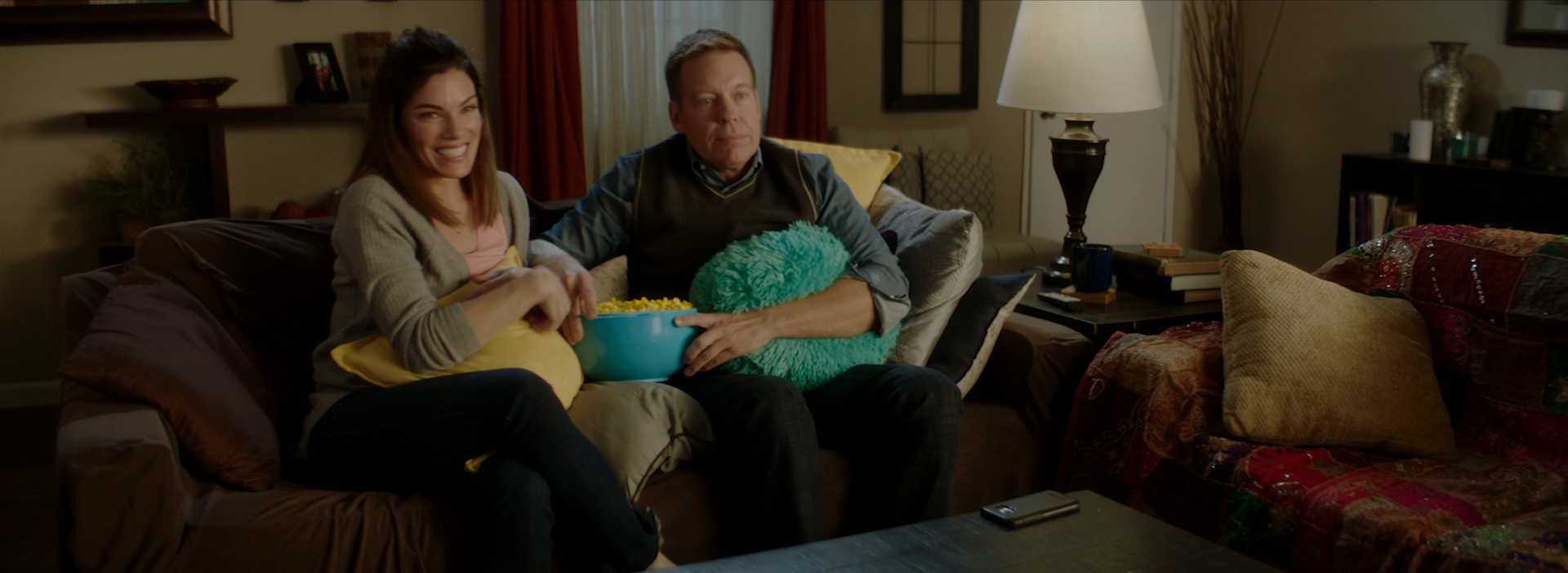 A woman and a man sitting on a brown couch in a living room, watching television. The woman is smiling and holding a yellow pillow, while the man is holding a bowl of popcorn and a fluffy teal pillow. The living room has various decorative pillows, a side table with a lamp, and framed pictures on the wall.