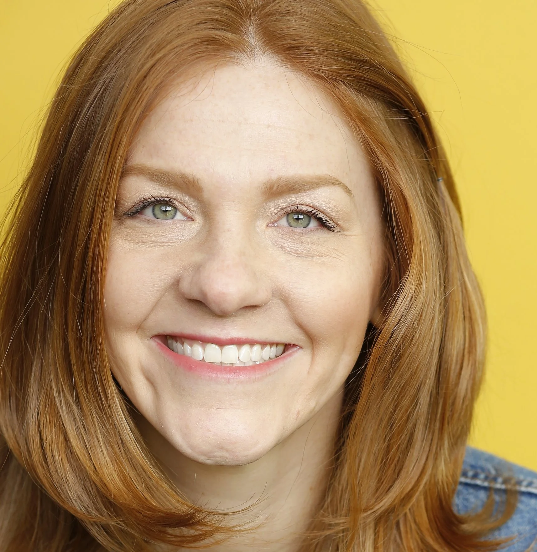 Close-up of a smiling woman with red hair and green eyes against a yellow background.