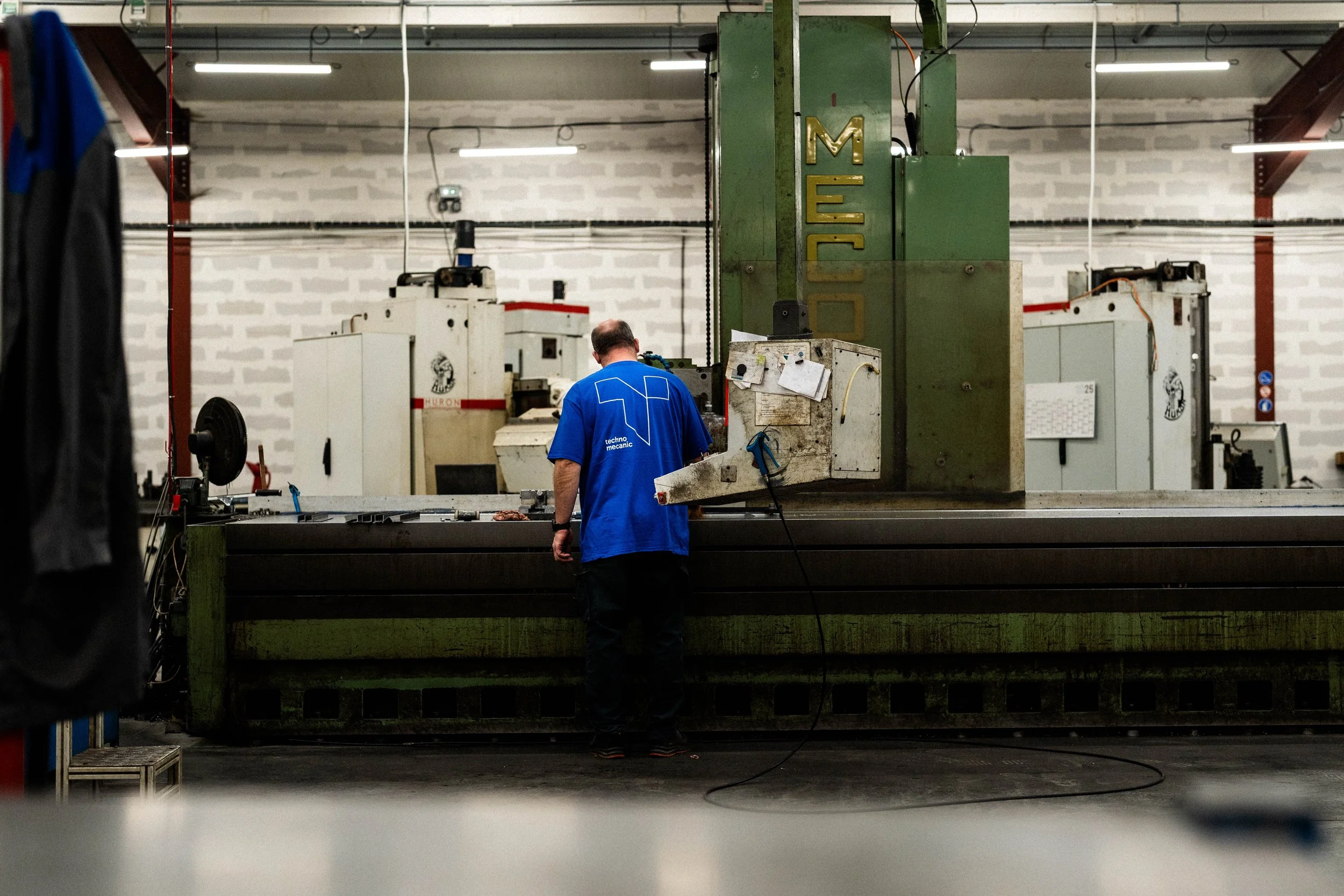 Un homme travaille dans une usine mécanique, regardant un grand équipement industriel vert avec le mot 'MECO' écrit dessus, dans une pièce éclairée par des lumières suspendues.