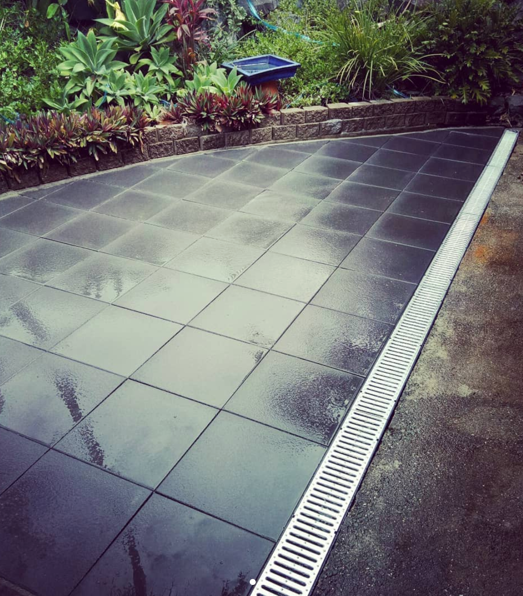 A wet black tiled surface outdoors, bordered by a metal drain grate along the edge, with lush green plants and a brick wall in the background. Designed by Romain Martens Stump Removal, Landscaping and Paving.