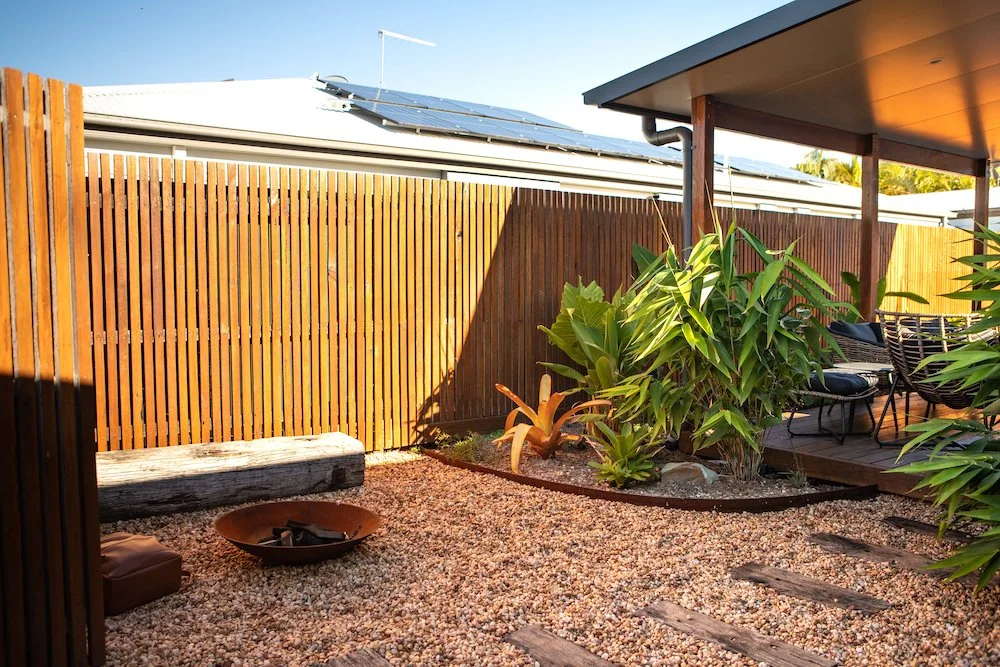Backyard patio with gravel ground, a large green plant, a wooden bench, patio furniture, a wooden fence, and solar panels on the roof. Designed by Romain Martens Stump Removal, Landscaping and Paving.