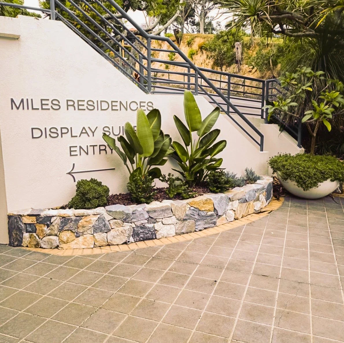 Sign indicating the entrance to Miles Residences, with a landscaped area featuring large green plants and a stone border, beside tiled ground and a staircase with black railing. Designed by Romain Martens Stump Removal, Landscaping and Paving.