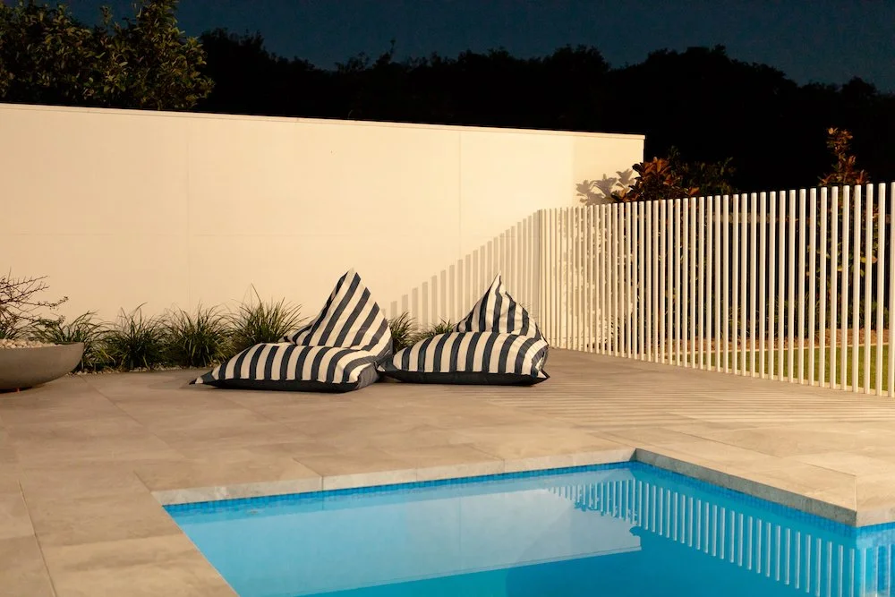 A modern outdoor pool area at dusk with two striped bean bag chairs on a tiled deck, surrounded by a white fence and plants, with a dark evening sky in the background. Designed by Romain Martens Stump Removal, Landscaping and Paving.