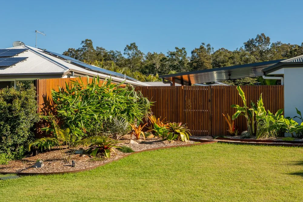 Residential backyard with a wooden fence, lush tropical plants, a green lawn, and solar panels on the roof in the background under a clear blue sky. Designed by Romain Martens Stump Removal, Landscaping and Paving.