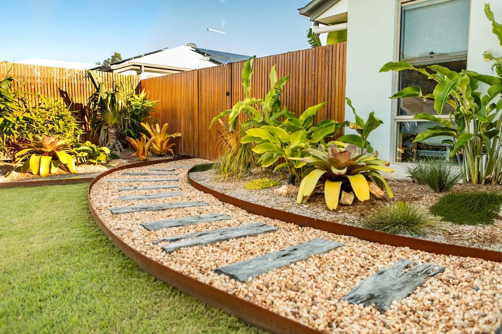 Backyard garden with a gravel pathway bordered by rusted metal edging, surrounded by various plants and a wooden fence, with a house and window on the right. Designed by Romain Martens Stump Removal, Landscaping and Paving.