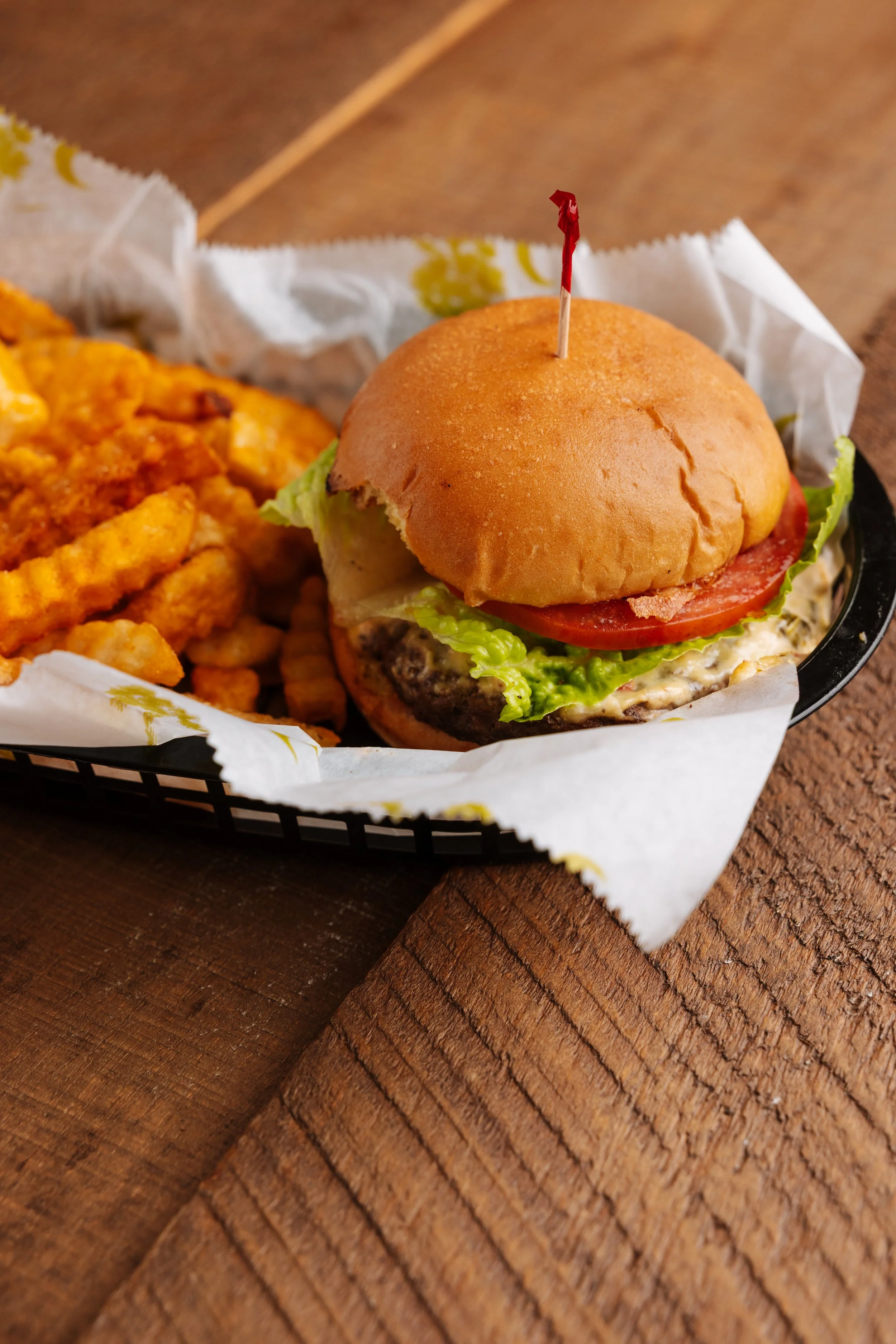 A cheeseburger with lettuce, tomato, and a pressed beef patty on a bun, served with curly fries in a black basket with paper liner on a wooden surface.