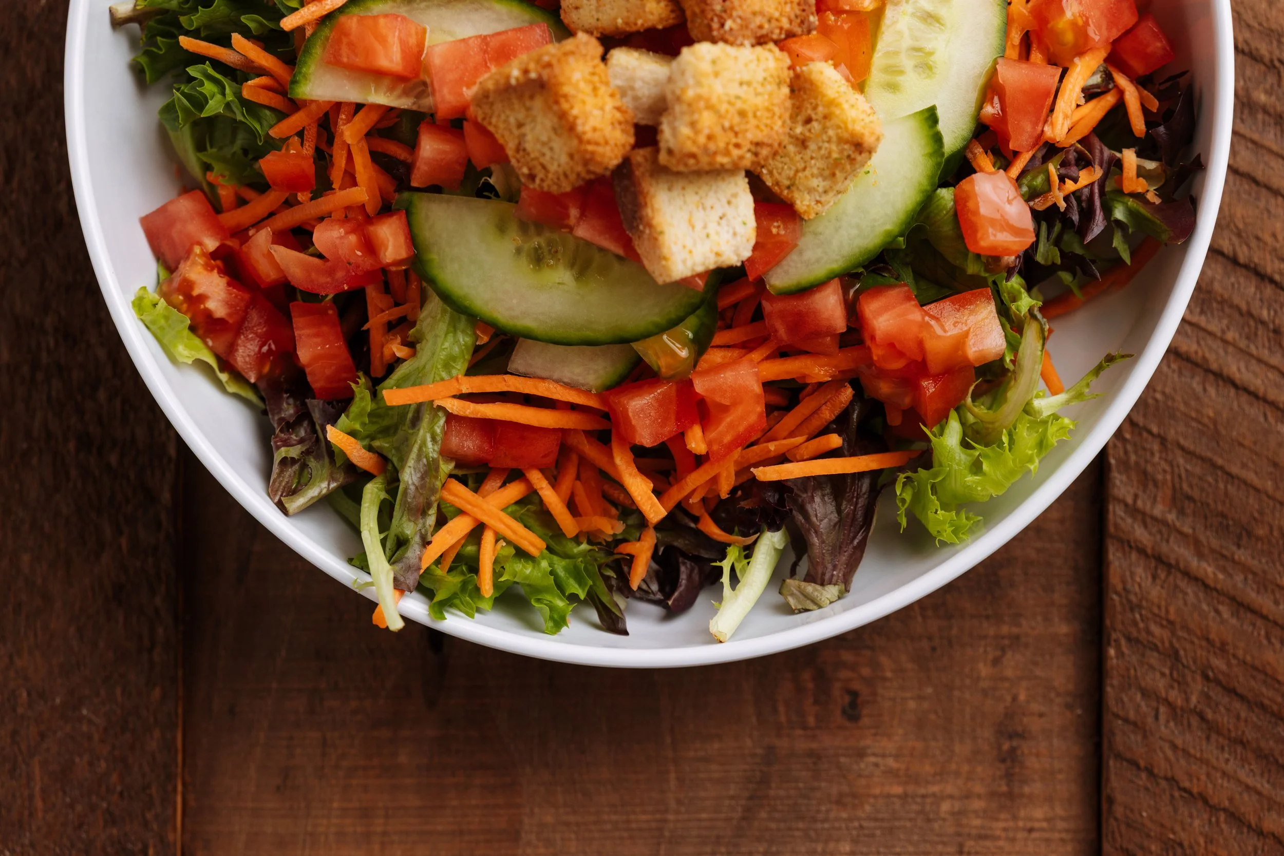 Close-up of a fresh vegetable salad in a white bowl, containing chopped lettuce, shredded carrots, diced tomatoes, cucumber slices, and croutons, placed on a wooden surface.