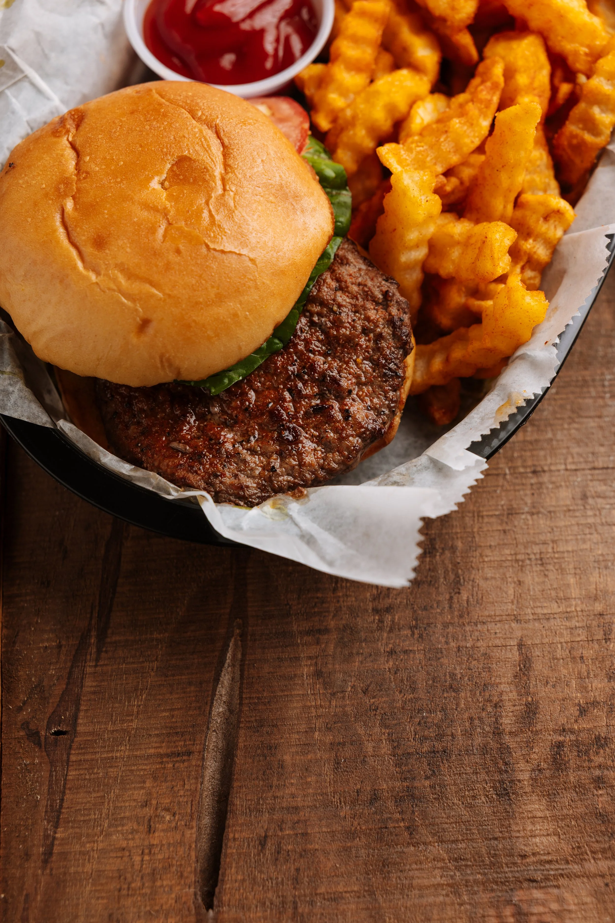 Close-up of a cheeseburger with lettuce in a bun, served with crinkle-cut French fries and a small cup of ketchup in a black basket lined with white paper, on a wooden table.