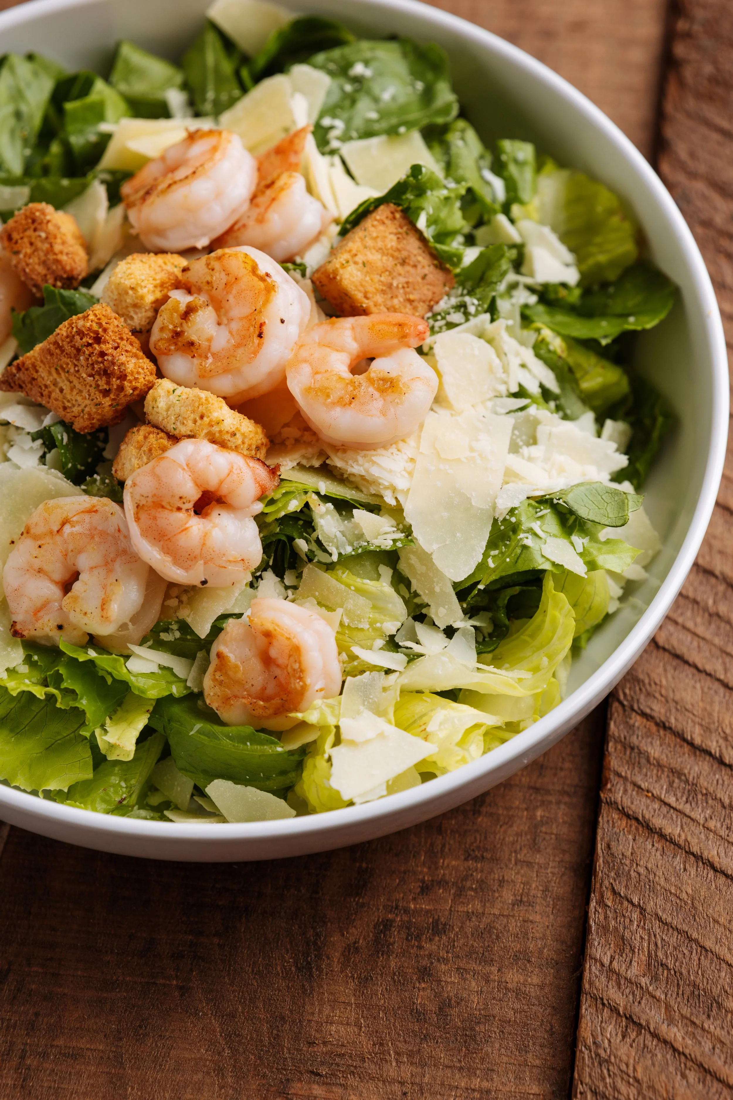 Caesar salad with shrimp, croutons, shredded cheese, and lettuce in a white bowl on a wooden surface.