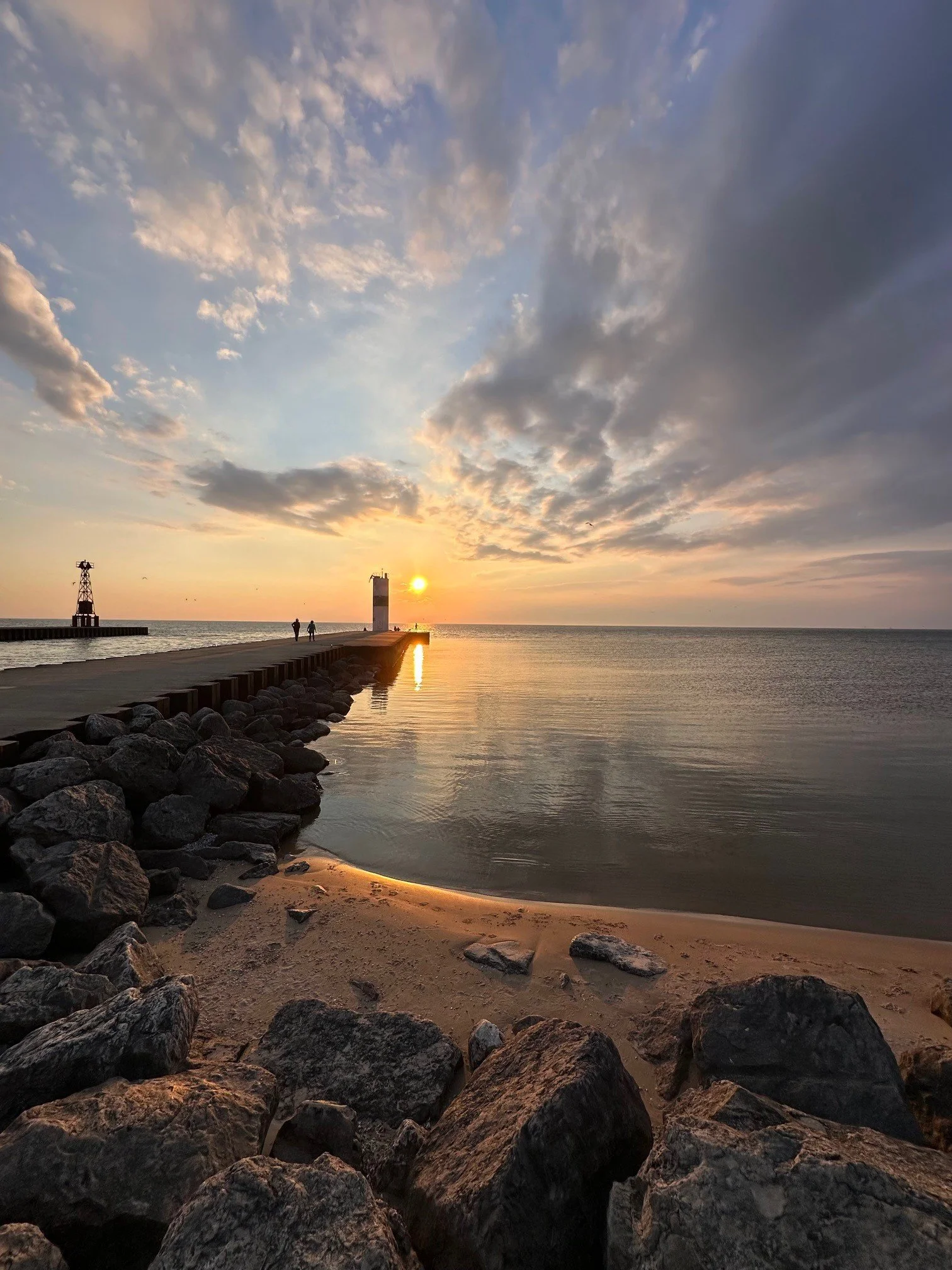 Sunset over a pier with two small buildings, rocks along the shoreline, and a partly cloudy sky.