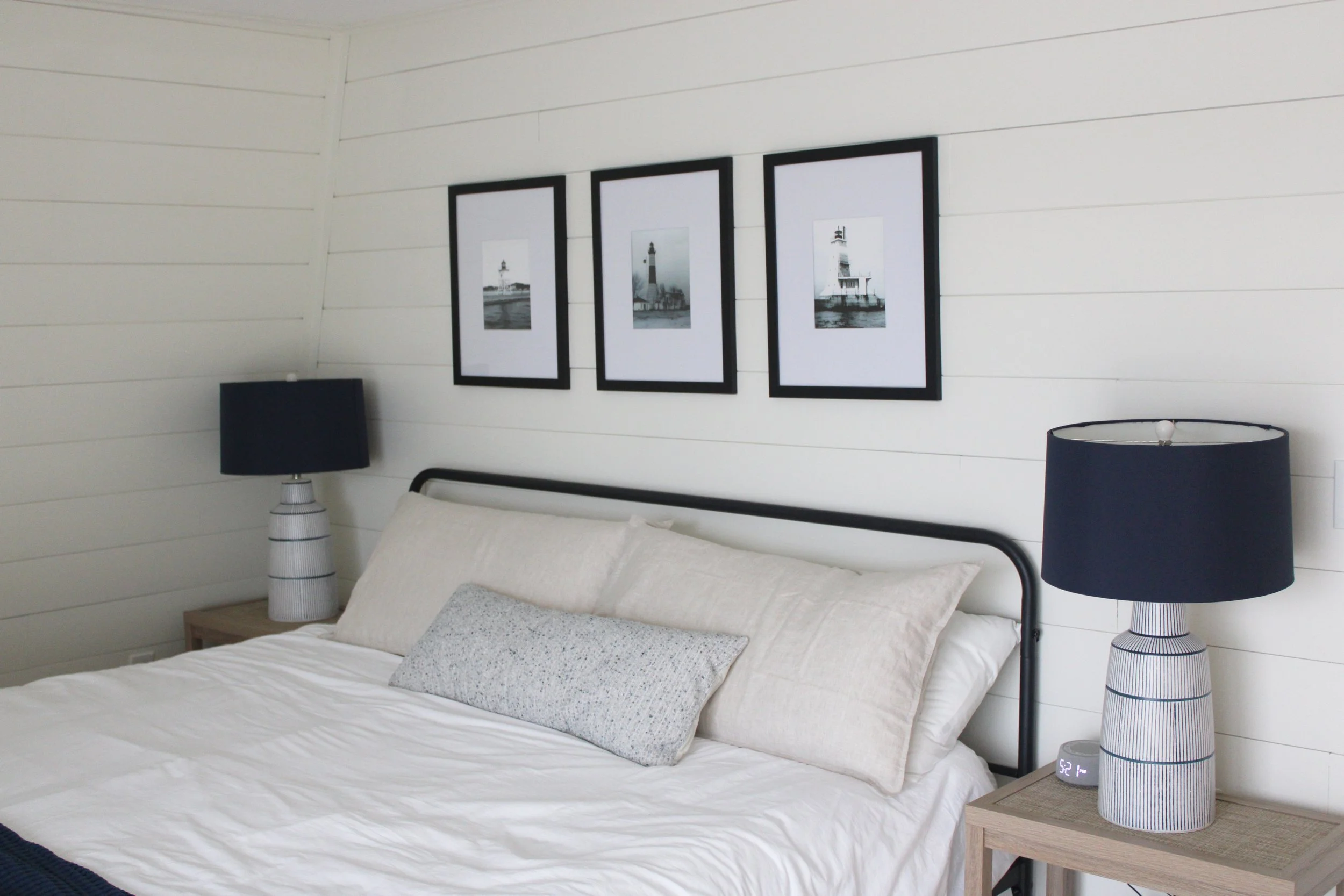 A bedroom with a bed, two nightstands with lamps, and three framed black and white lighthouse pictures on the wall.