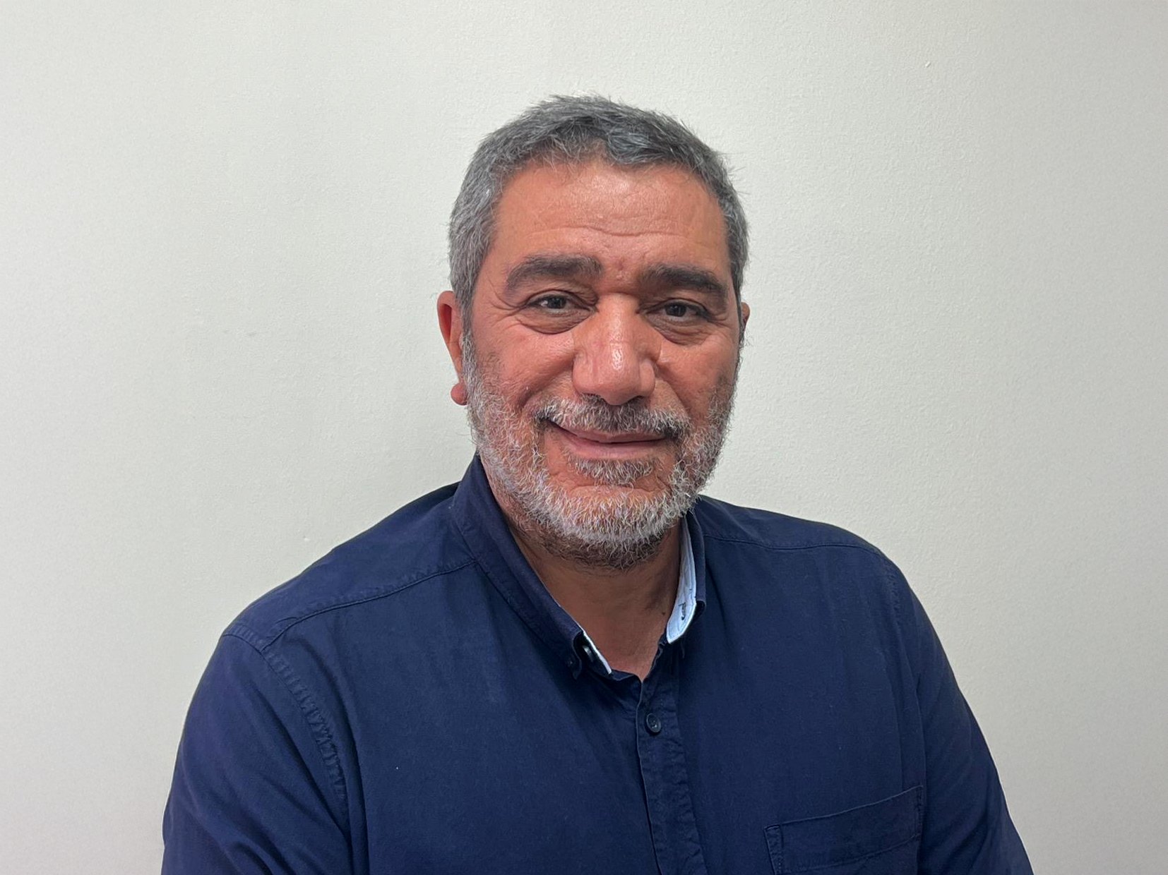 A middle-aged man with gray hair and beard, wearing a dark blue collared shirt, smiling in front of a plain light-colored wall.