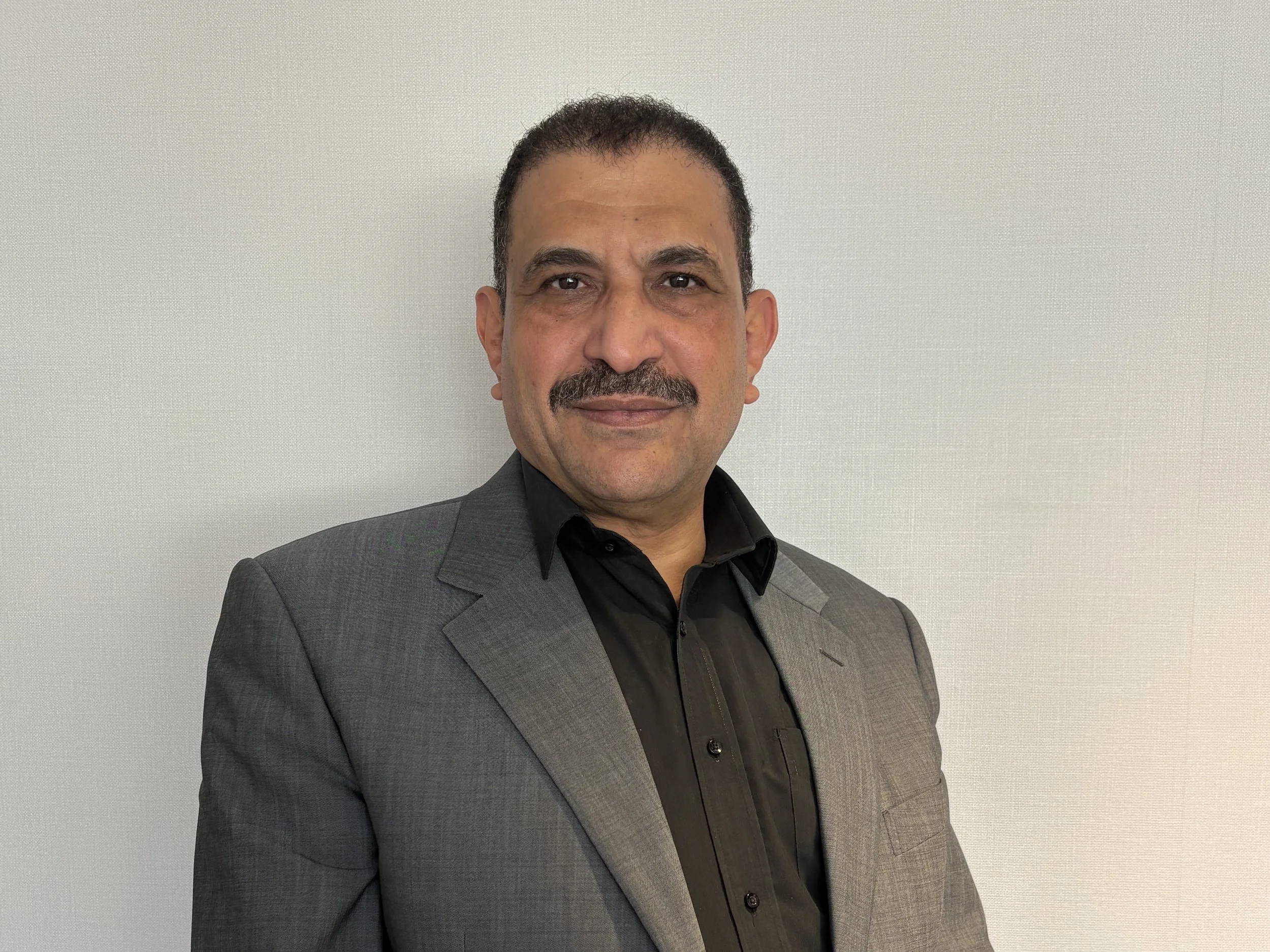 A man with short curly hair, a mustache, and medium skin tone, wearing a gray suit jacket and a black shirt, posing against a plain off-white wall.