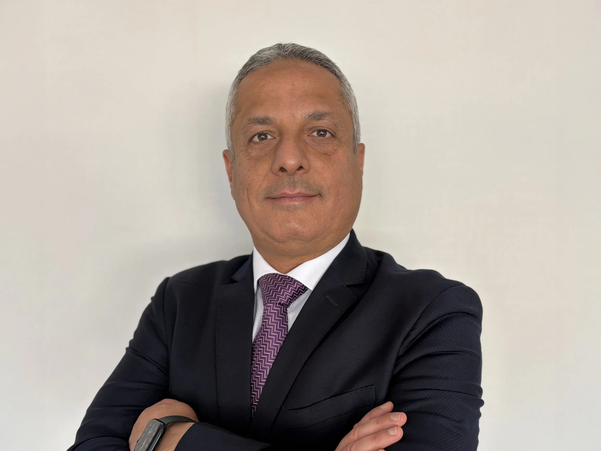 A professional man in a dark suit with a patterned purple tie, standing with arms crossed against a plain light-colored background.