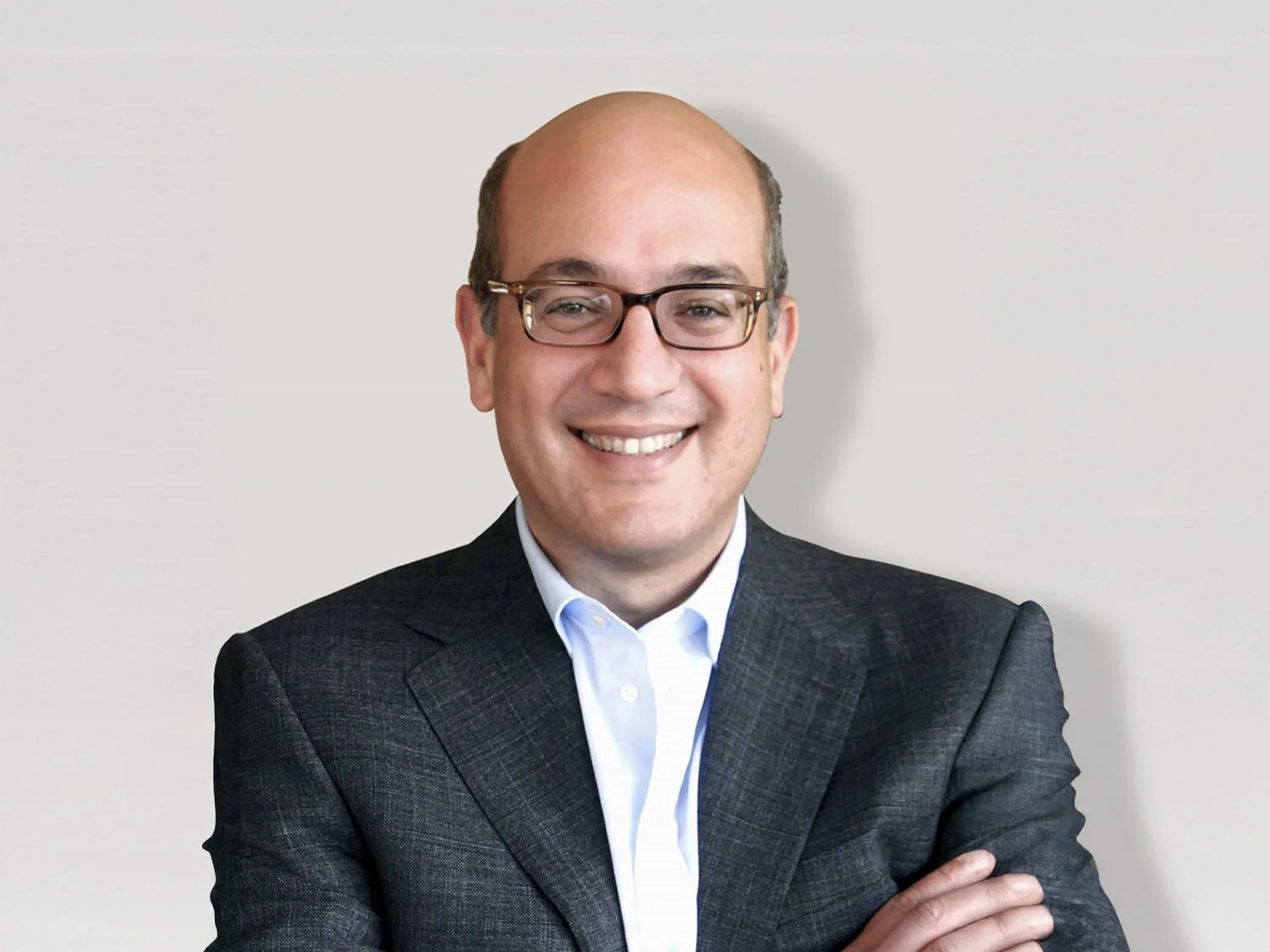 Smiling man in a suit with glasses, standing against a plain background.