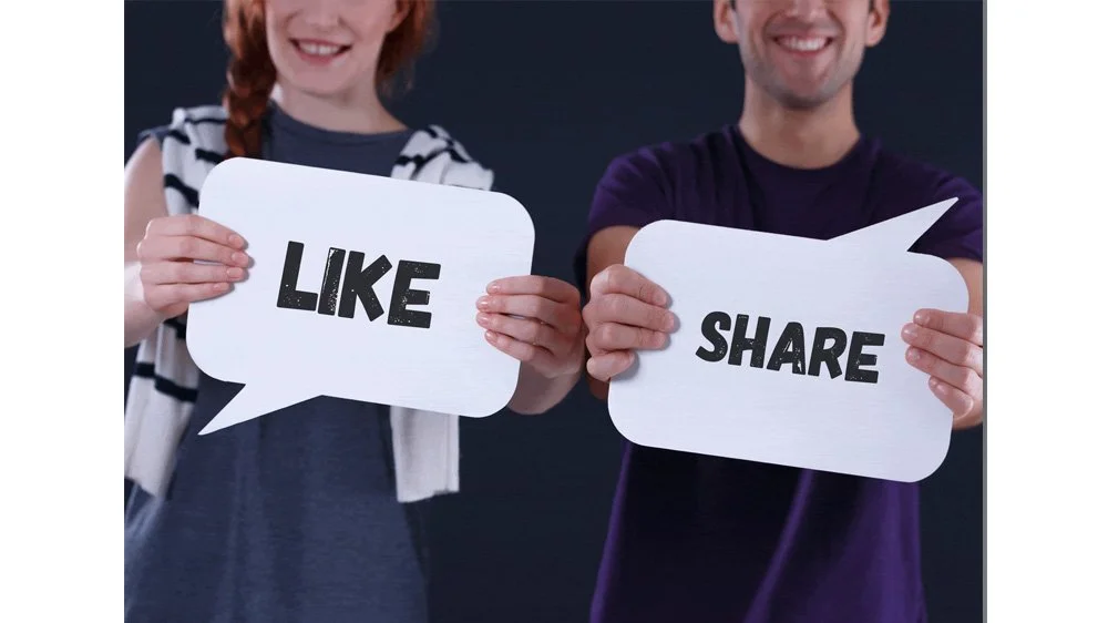 Two people, a woman with red hair and a man, smiling and holding speech bubbles that say "LIKE" and "SHARE"