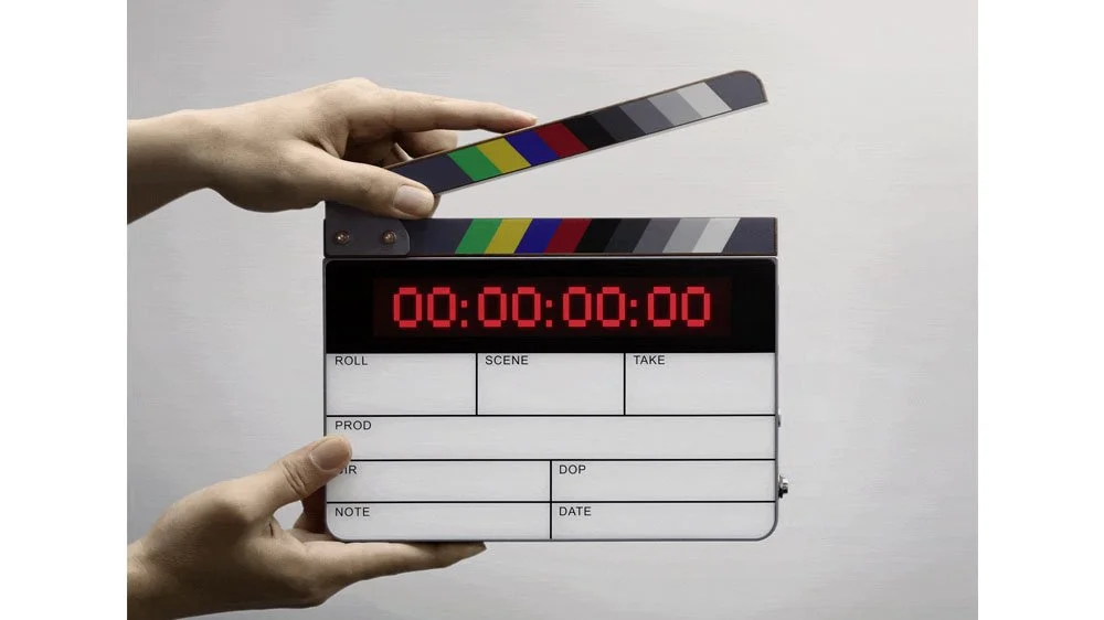 A person holding a film slate with a digital timer display showing 00:00:00:00, and the background is plain white.