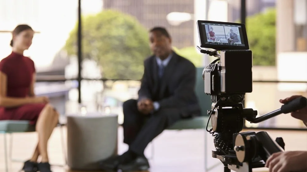 Two business people sat on a sofa being filmed by a camera
