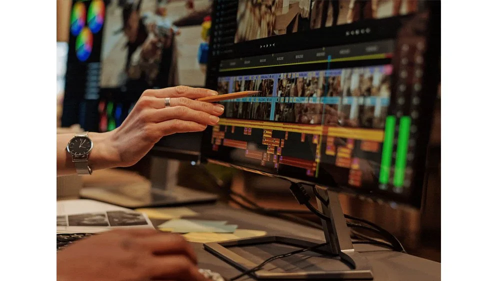 Person using a pencil to edit a video timeline on a computer monitor in a workspace.