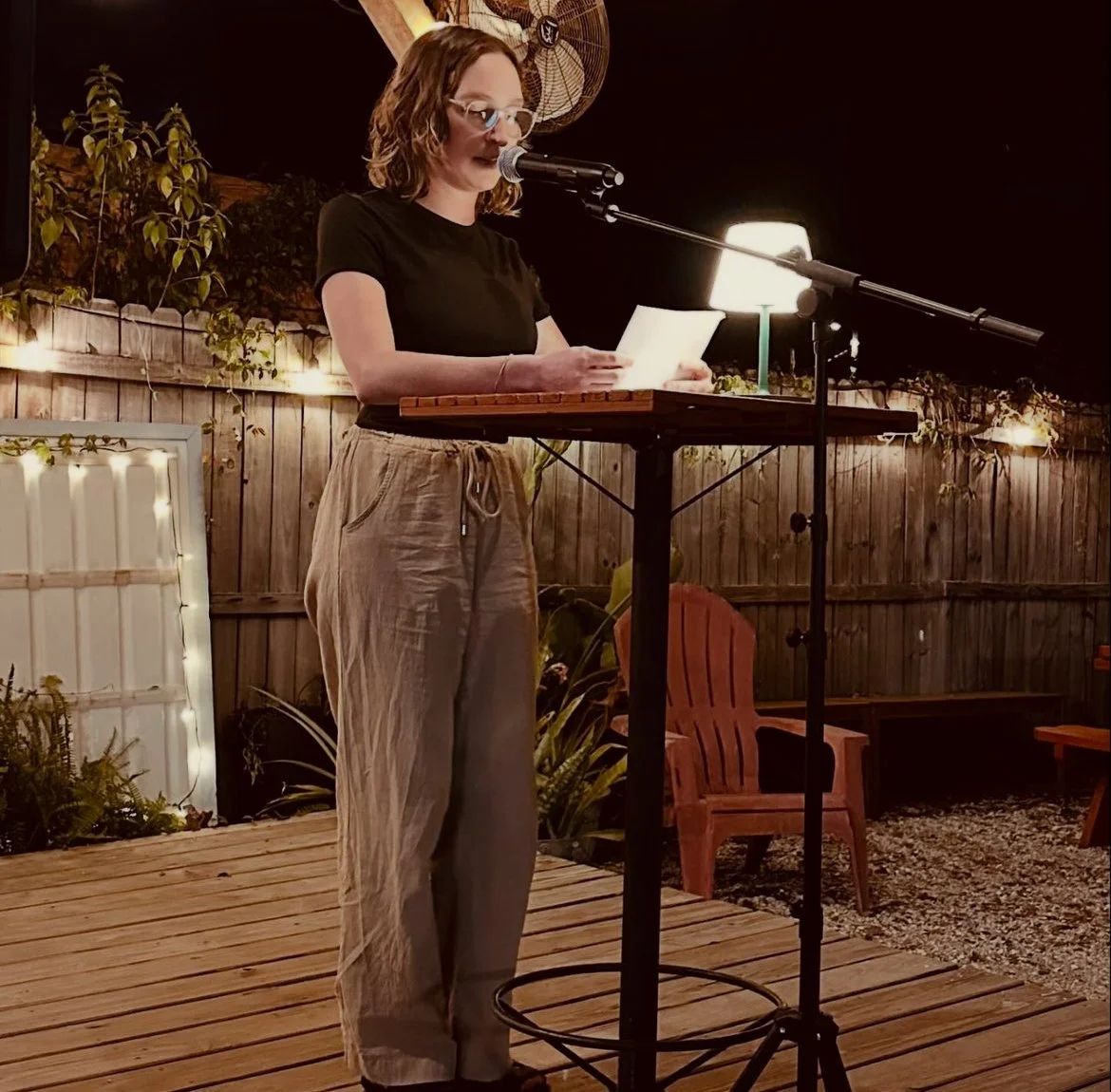 Mary Shaver wearing a black shirt and beige pants, standing outdoors at night, reading from papers into a microphone on a stand. There are string lights, a wooden fence, plants, and outdoor chairs in the background.