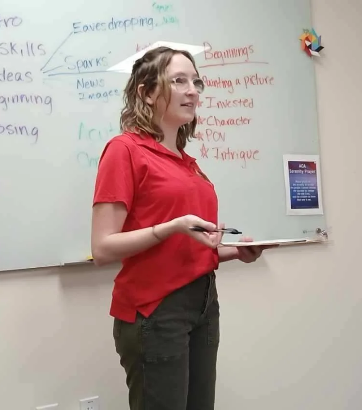 Mary Shaver wearing glasses and a red shirt, stands in front of a whiteboard with notes and a poster, holding a notebook.