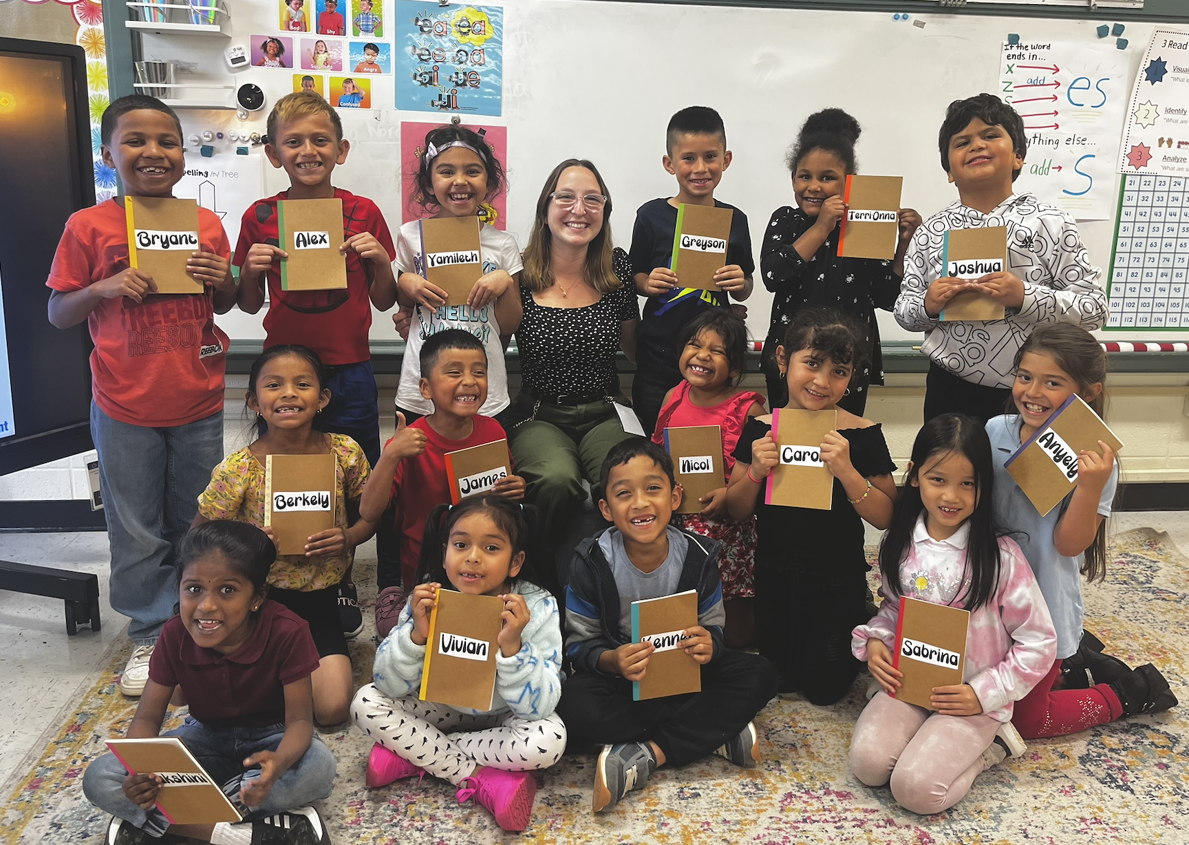 A classroom with a teacher (Mary Shaver) and 14 children. The children are holding notebooks with their names on them, smiling. The classroom has colorful posters and educational materials on the walls.