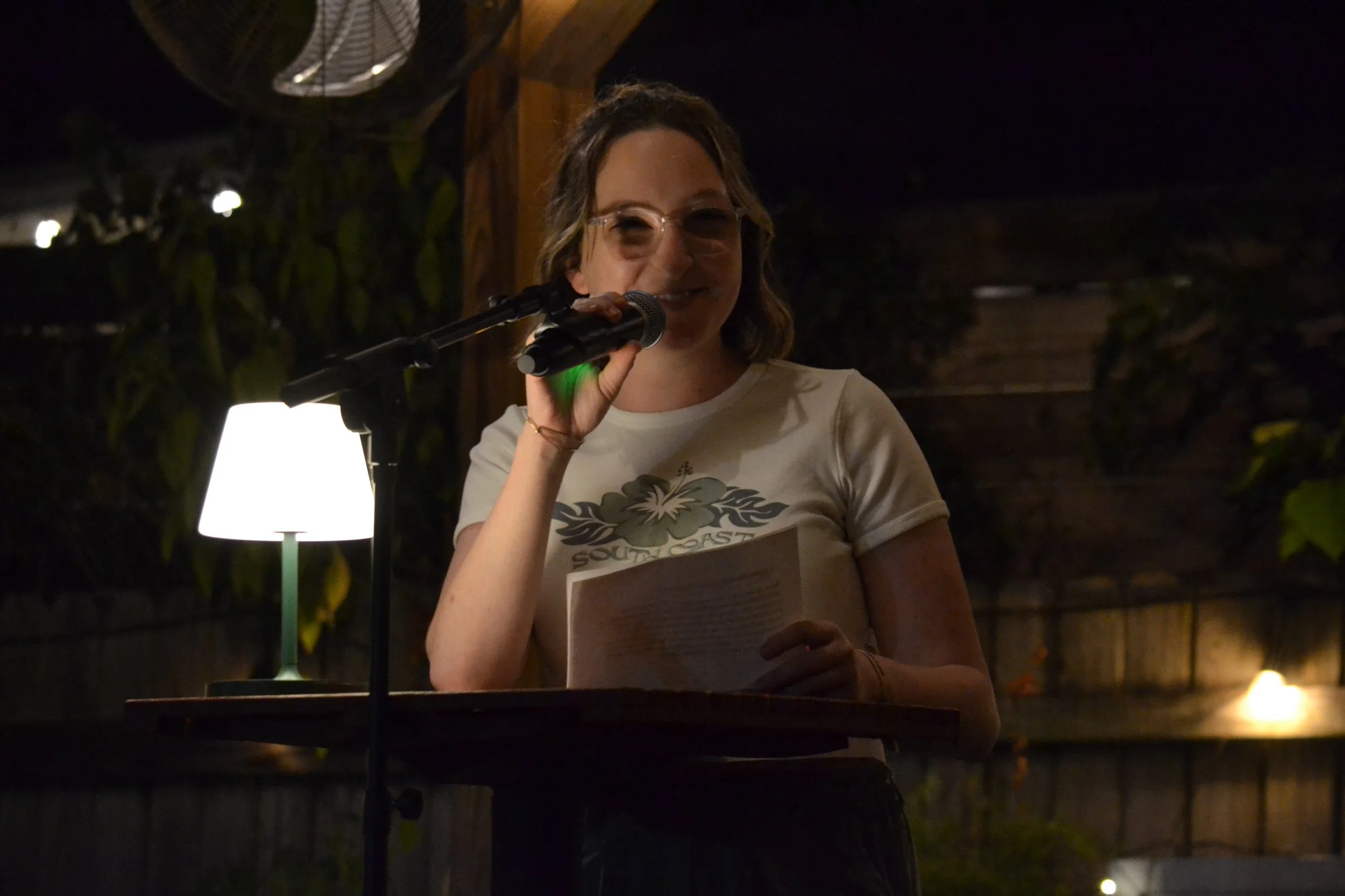 Mary Shaver standing at a small table, holding a microphone, speaking or reading, with a paper in her hand, in a dimly lit outdoor setting at night.