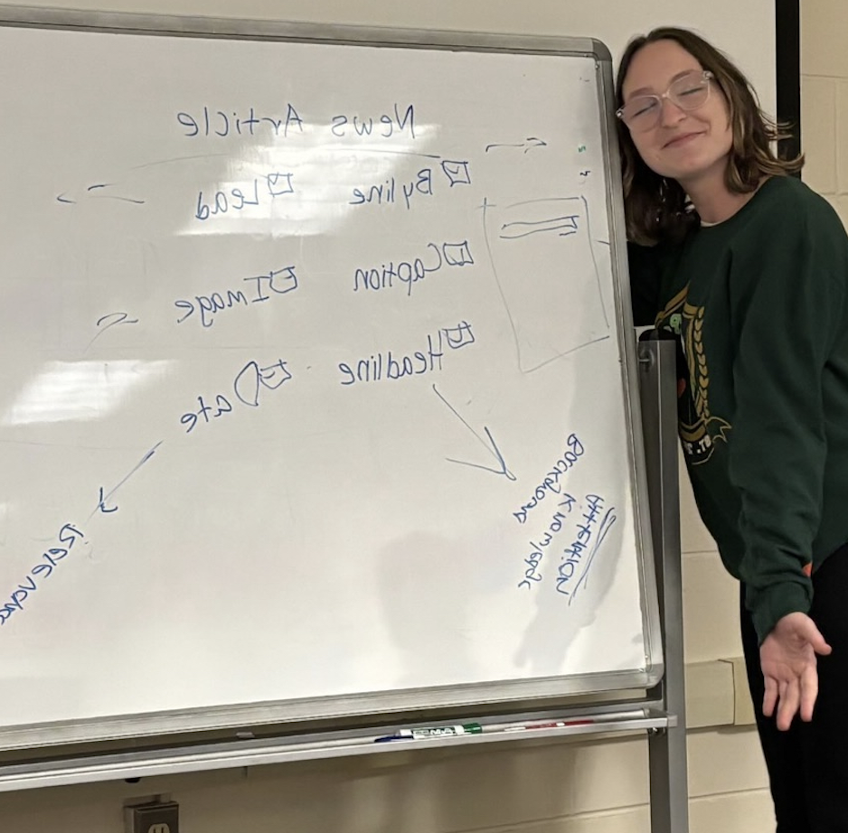 Mary Shaver smiling while standing next to a whiteboard that has handwritten notes and diagrams about composition.