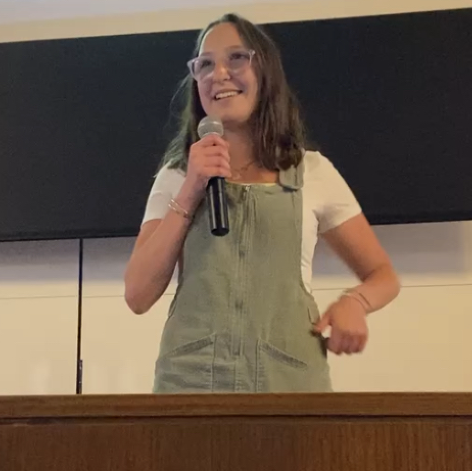 Mary Shaver with glasses and shoulder-length wavy brown hair is smiling and speaking into a microphone. She is wearing a light-colored shirt under olive green overalls and standing in front of a large dark screen or board.