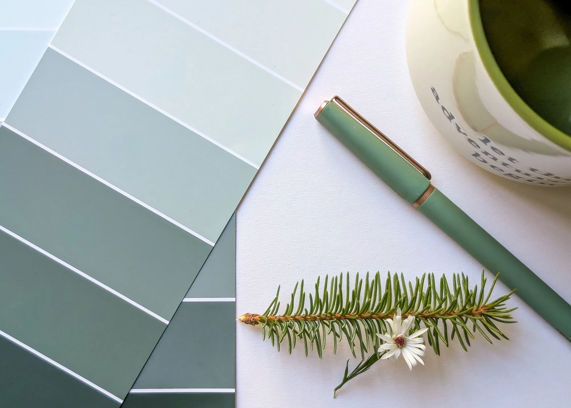 Color palette with shades of green, a green pen, a potted plant with dark green interior, a white flower with green leaves, and a white surface background.