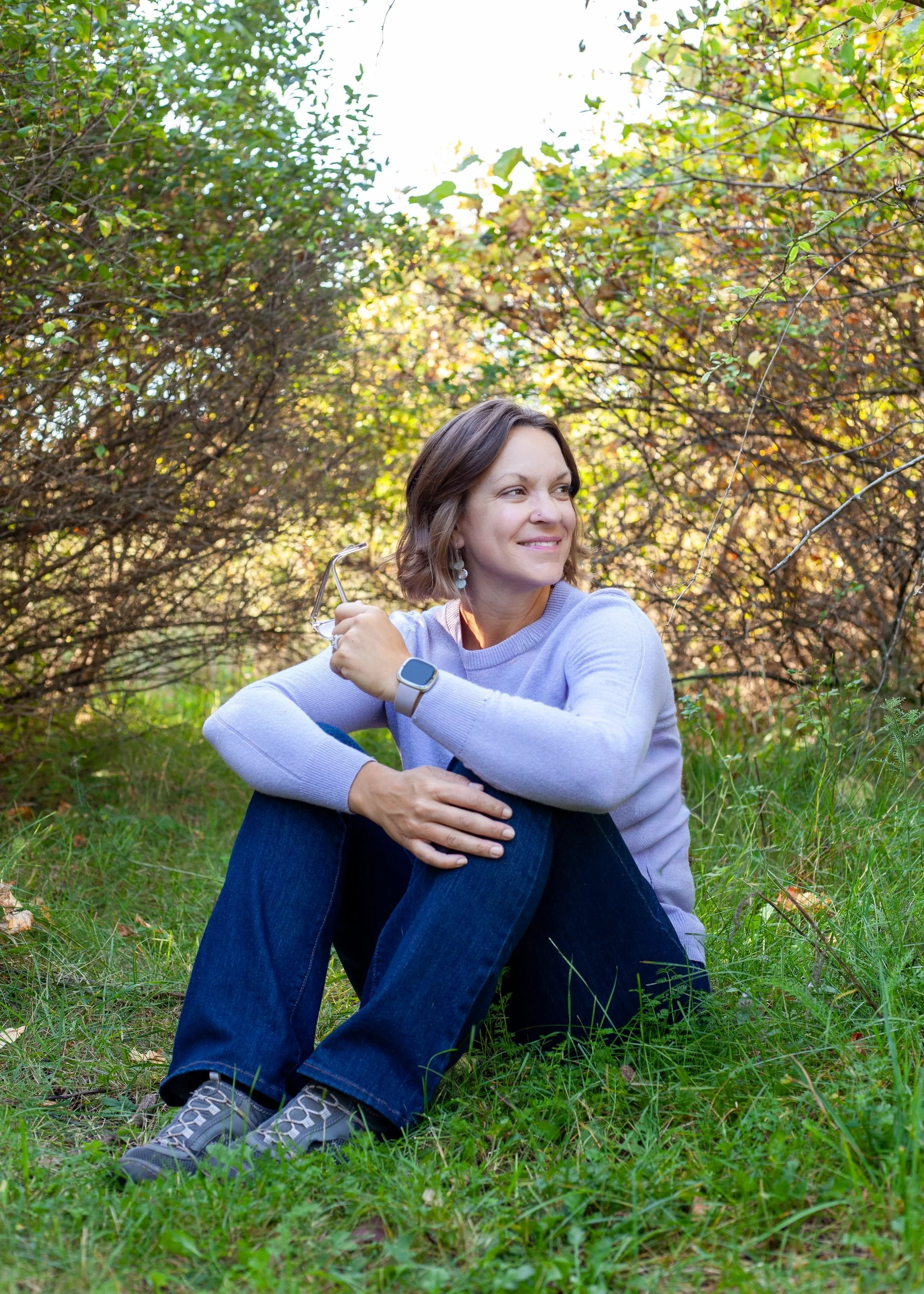 A woman sitting on the grass in a wooded area, smiling and relaxing, wearing a lavender sweater, dark jeans, and hiking shoes, holding glasses with trees in the background.