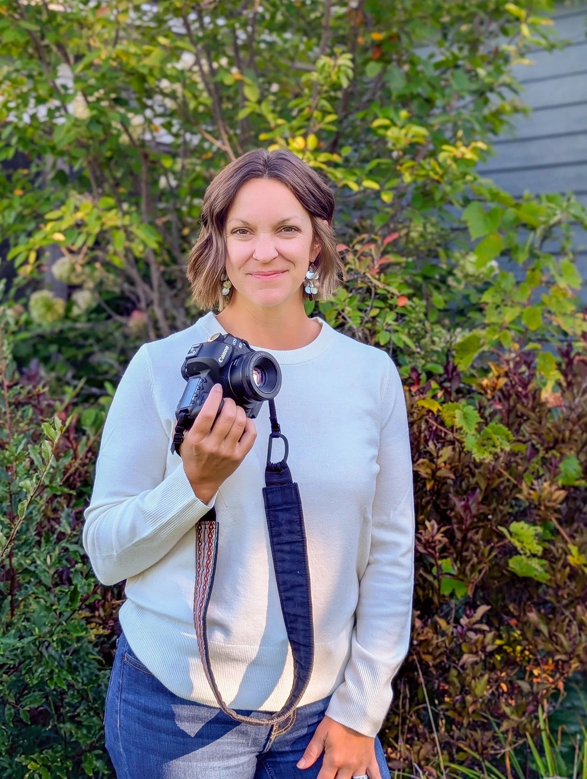 A woman holding a camera outdoors in front of green foliage and a house