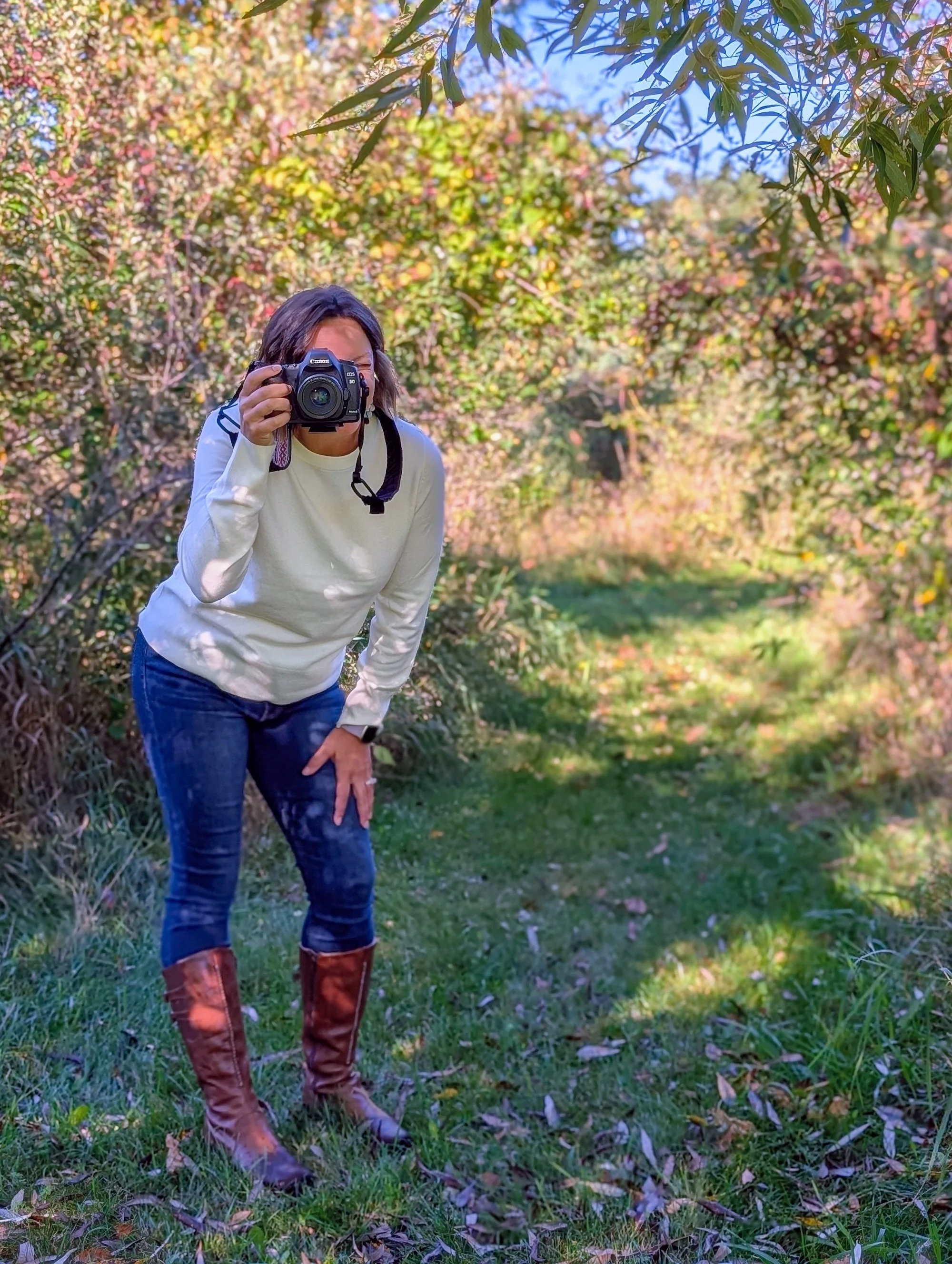 A woman in an outdoor setting, holding a camera up to her face, ready to take a photograph. She is wearing a white long-sleeve shirt, blue jeans, and brown knee-high boots, with a forested background.