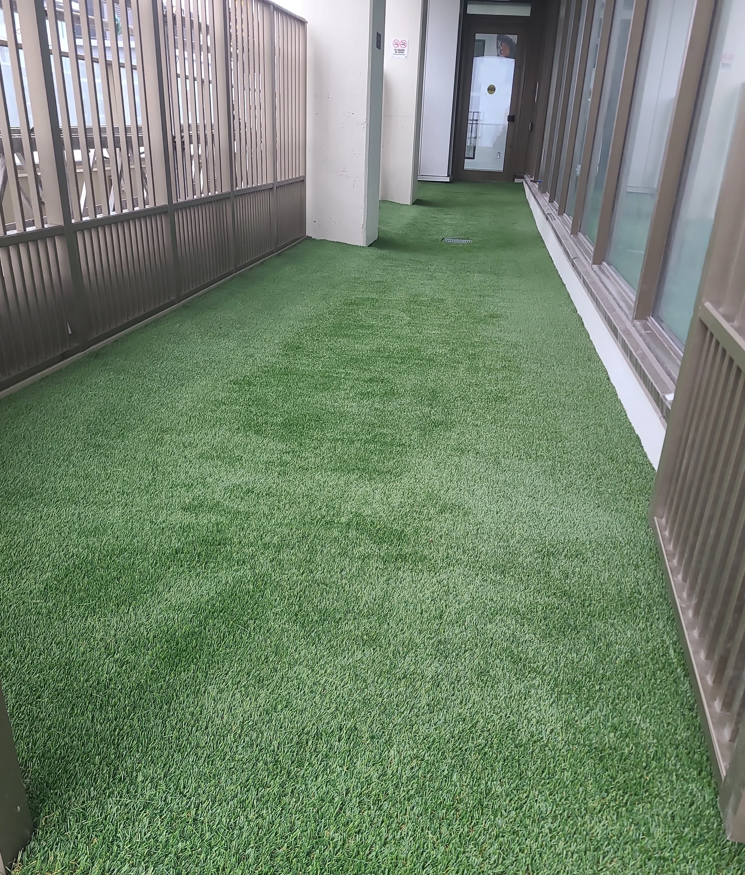 Empty balcony or walkway with green artificial grass flooring, metal railing on the left, glass windows on the right, and a door at the far end.