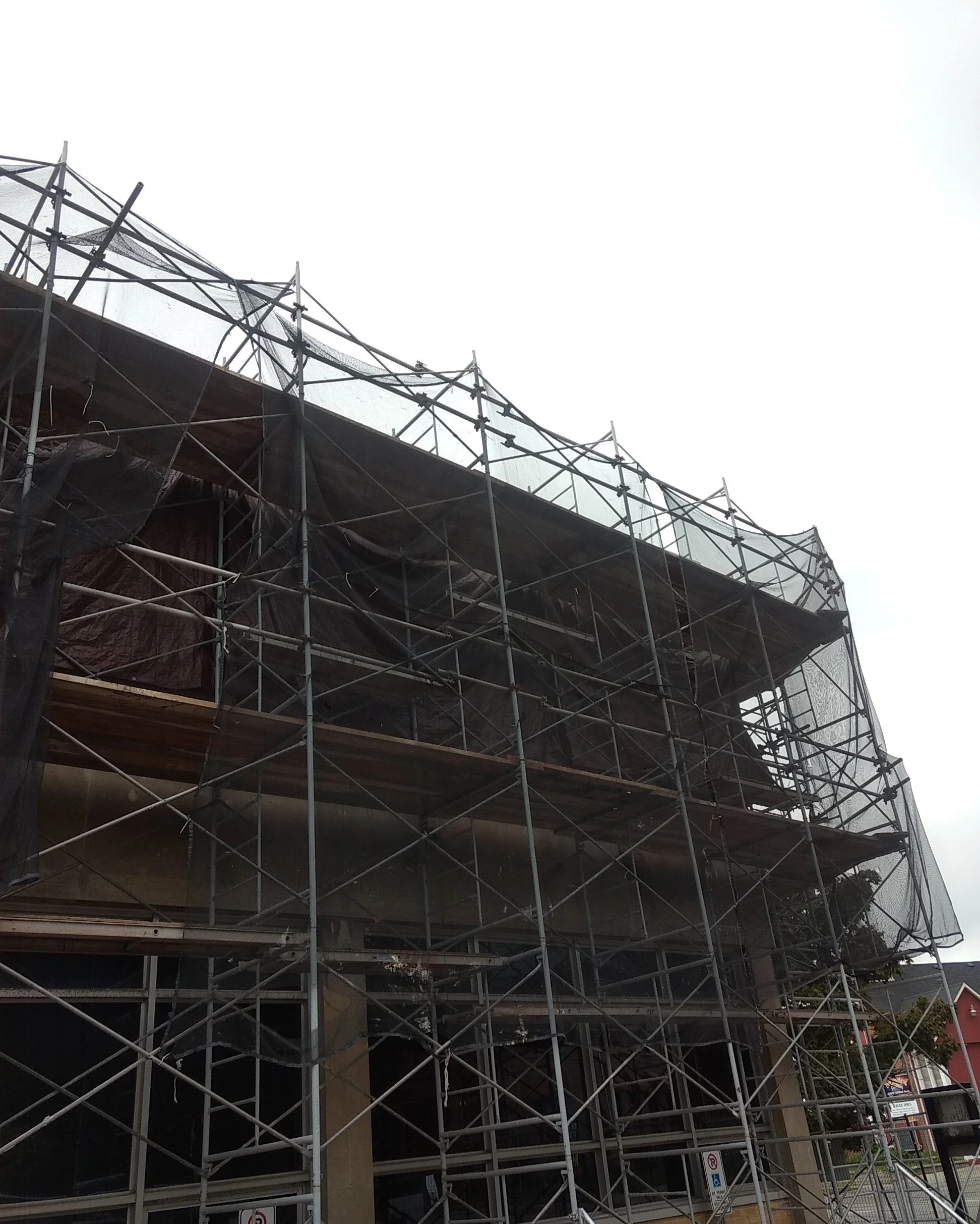 A building under construction with scaffolding and safety netting on all sides, set against an overcast sky.