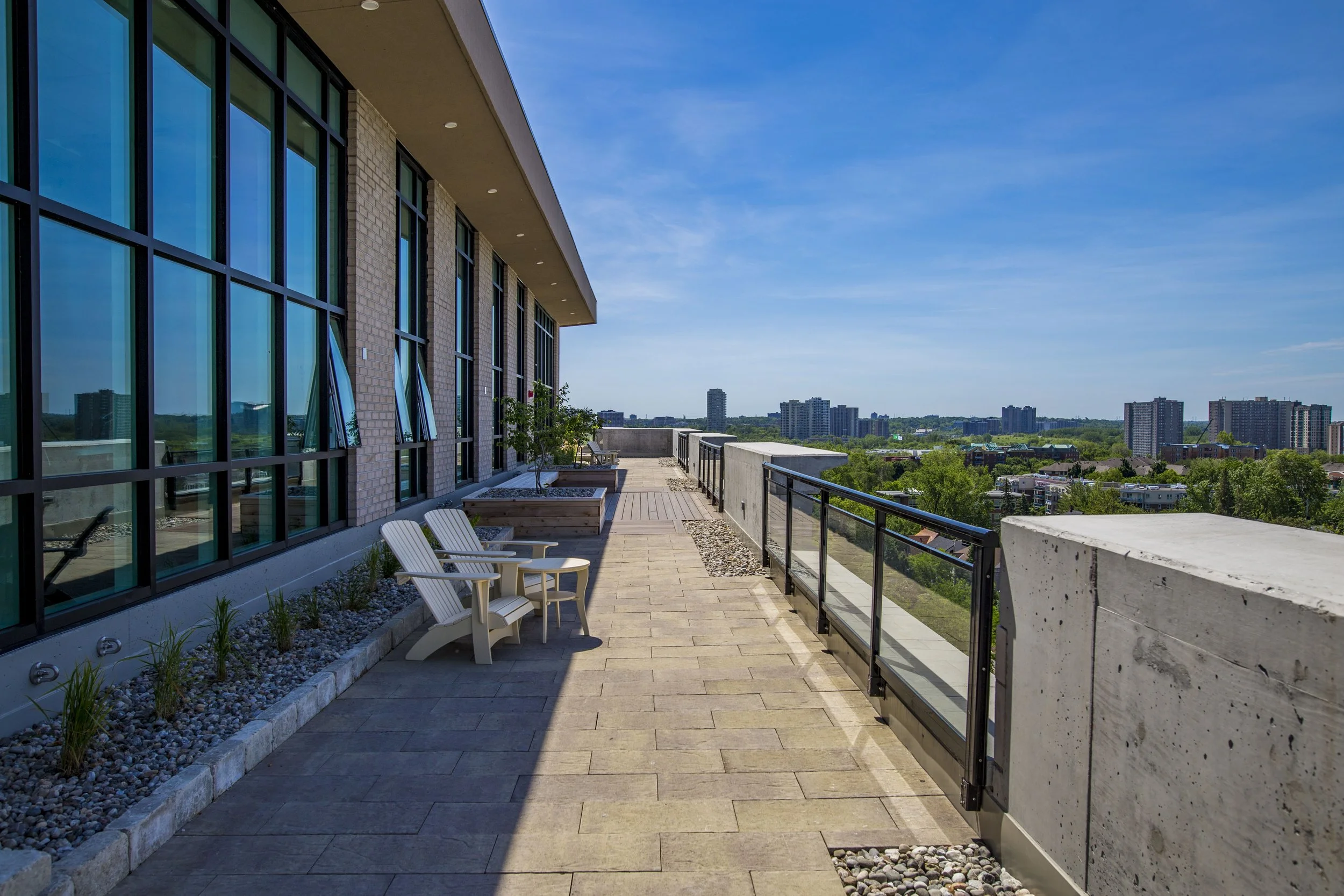 A spacious rooftop terrace with beige pavers, outdoor chairs, potted trees, and a glass railing overlooking a cityscape with high-rise buildings and green trees under a blue sky.