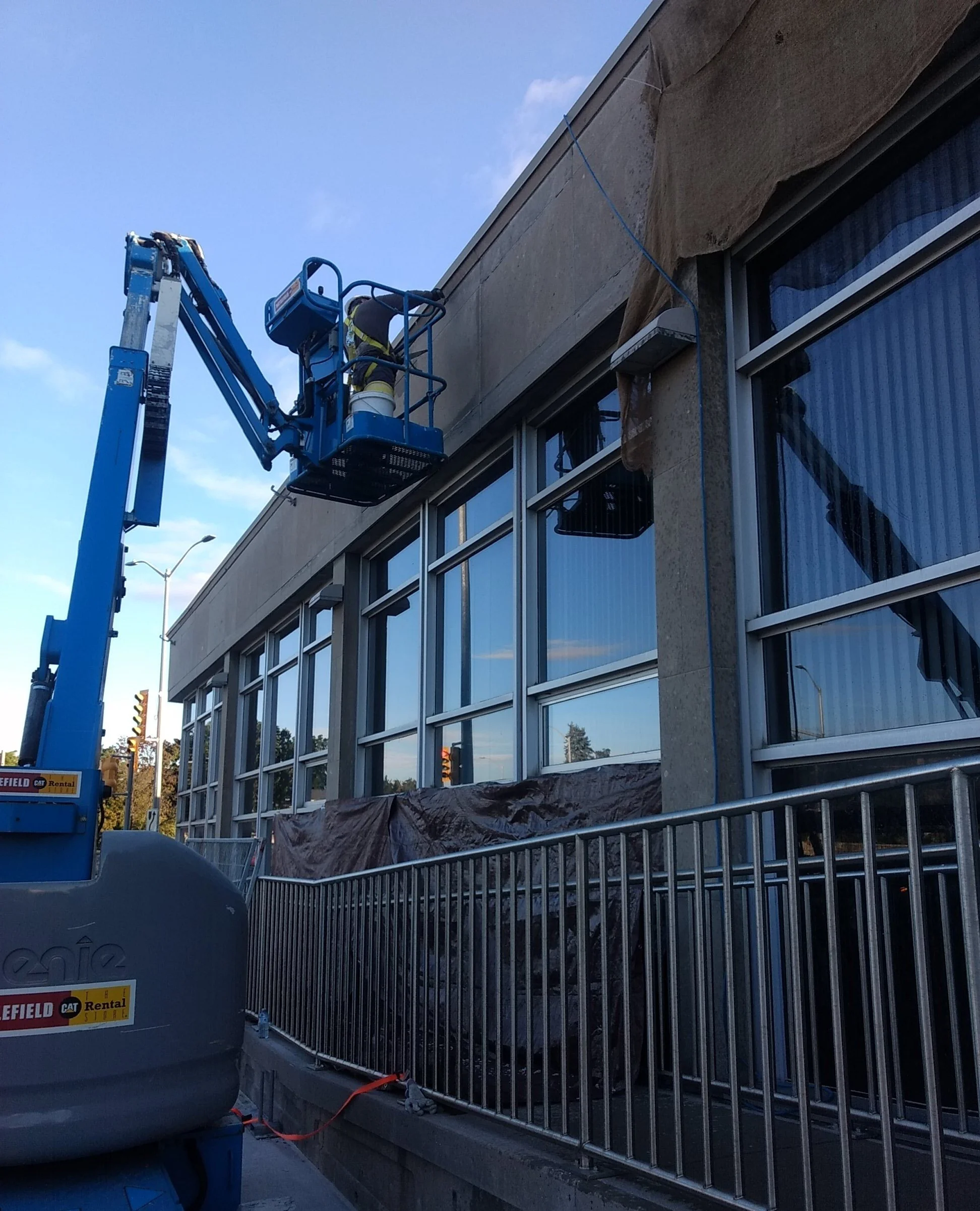 A worker on a blue cherry picker working on the exterior of a building with large glass windows, partially covered with brown material, during daytime.