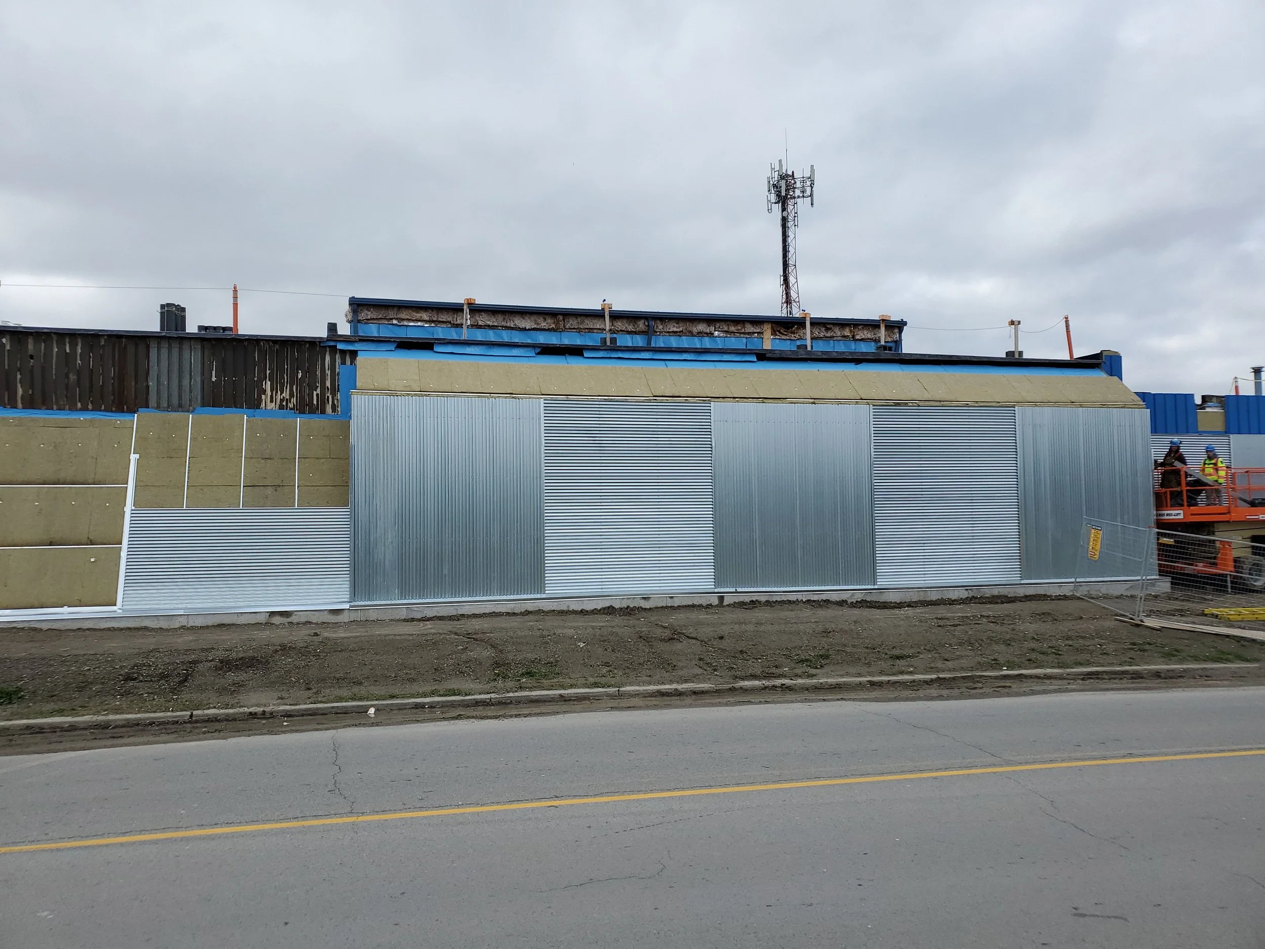 Construction site with metal panels and insulation on a building, construction workers on a lift, overcast sky.