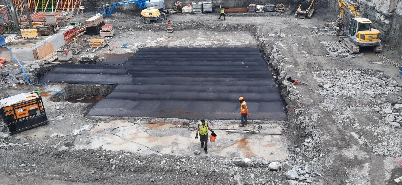 Construction site with workers and machinery, with a recently paved asphalt section in the center, surrounded by dirt and rocks.