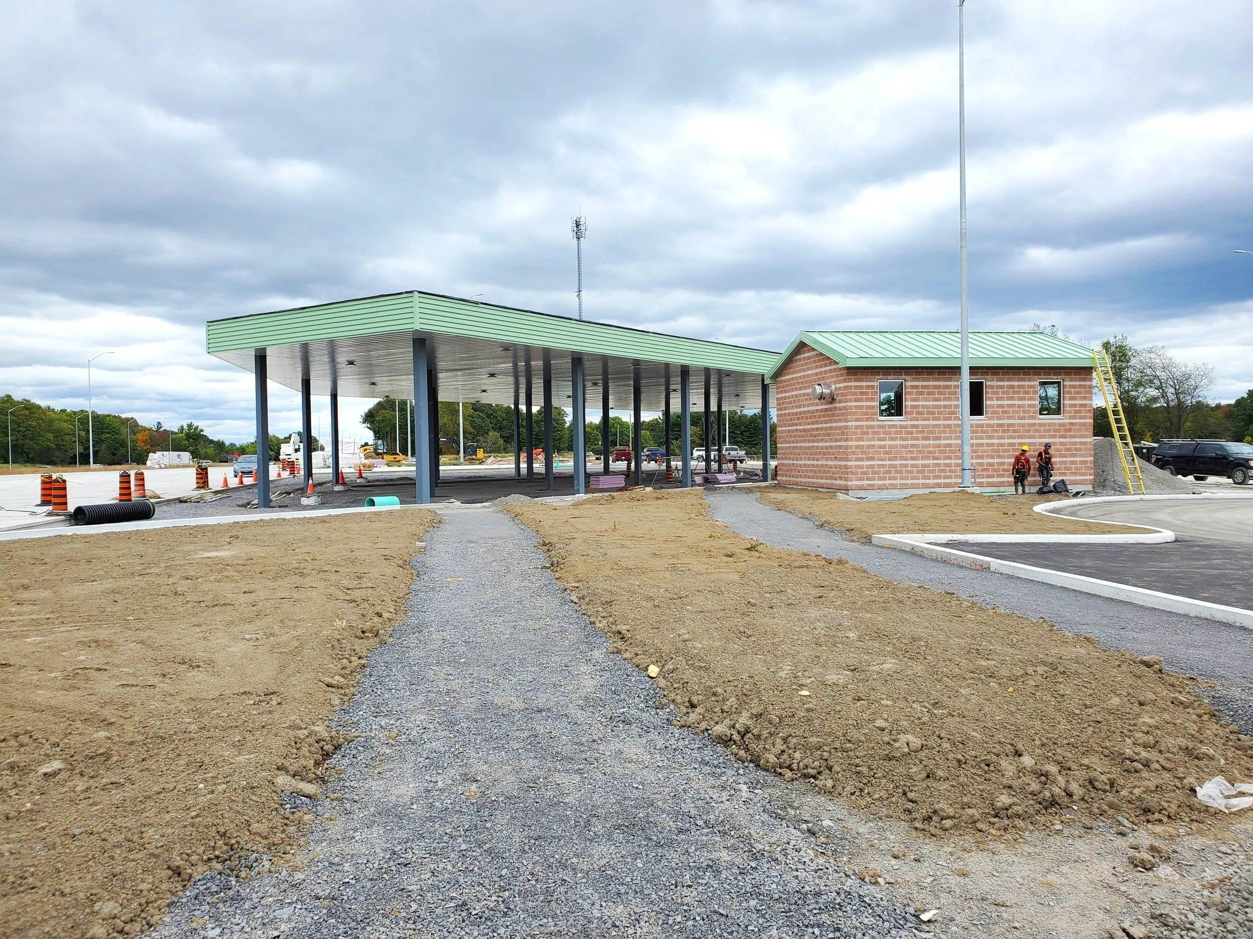 Construction site of a gas station or convenience store with a green metal roof, supported by multiple black columns. There are two workers standing near a small brick building with a green metal roof, ladder, and ventilation pipe. The foreground has