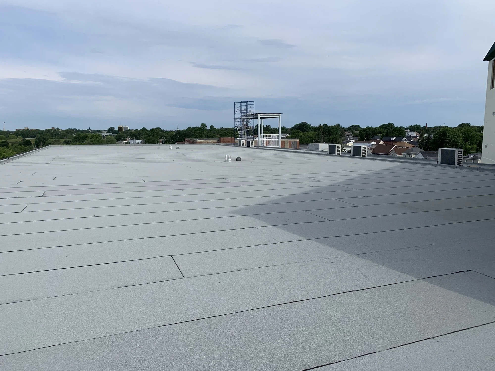 A view of rooftops and treetops from a building with a gray metal roof, featuring rooftop vents, a tower, and a sky with scattered clouds.