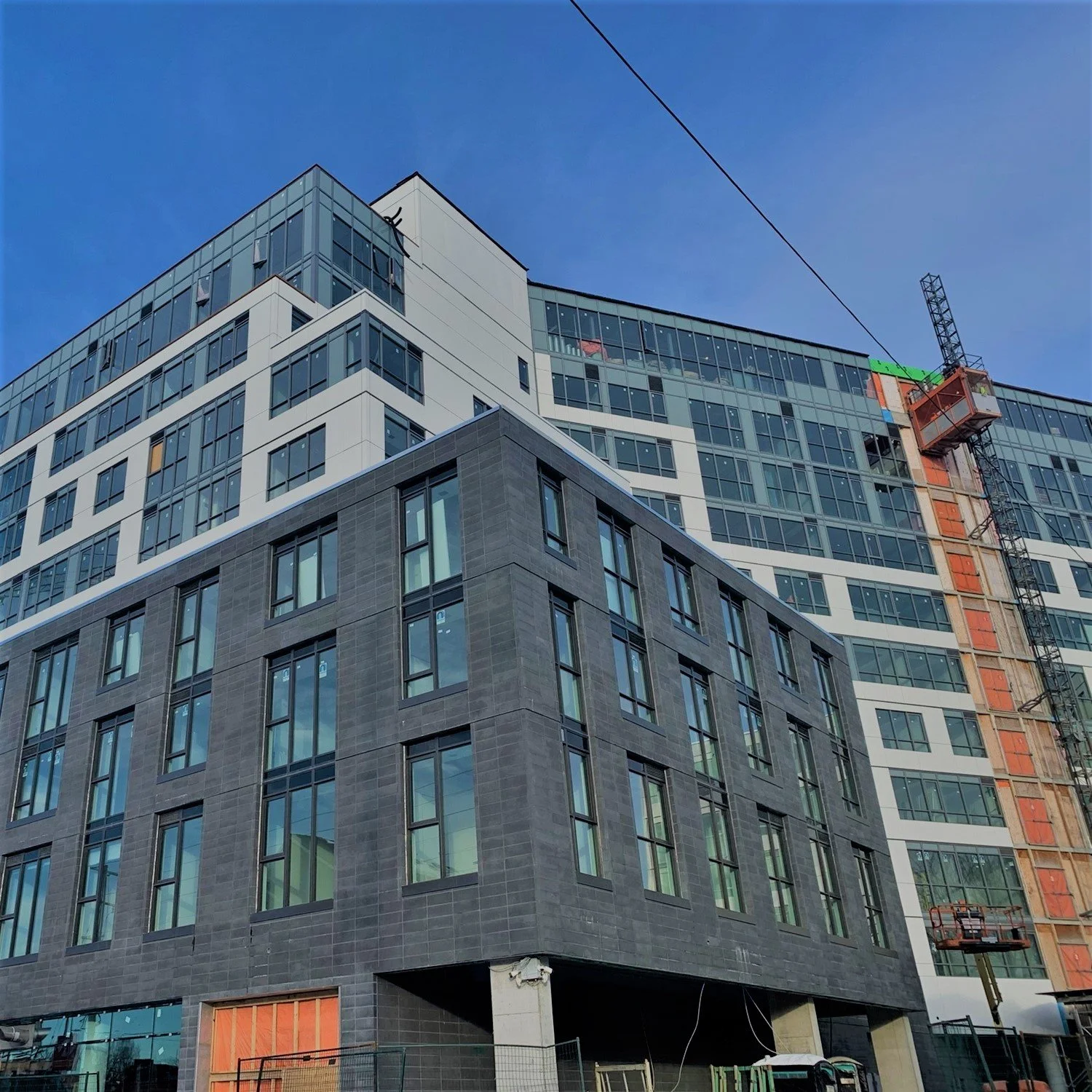 A modern multi-story building under construction with a dark gray brick facade and large glass windows. A construction lift is working on the upper floors, and the sky is clear and blue.
